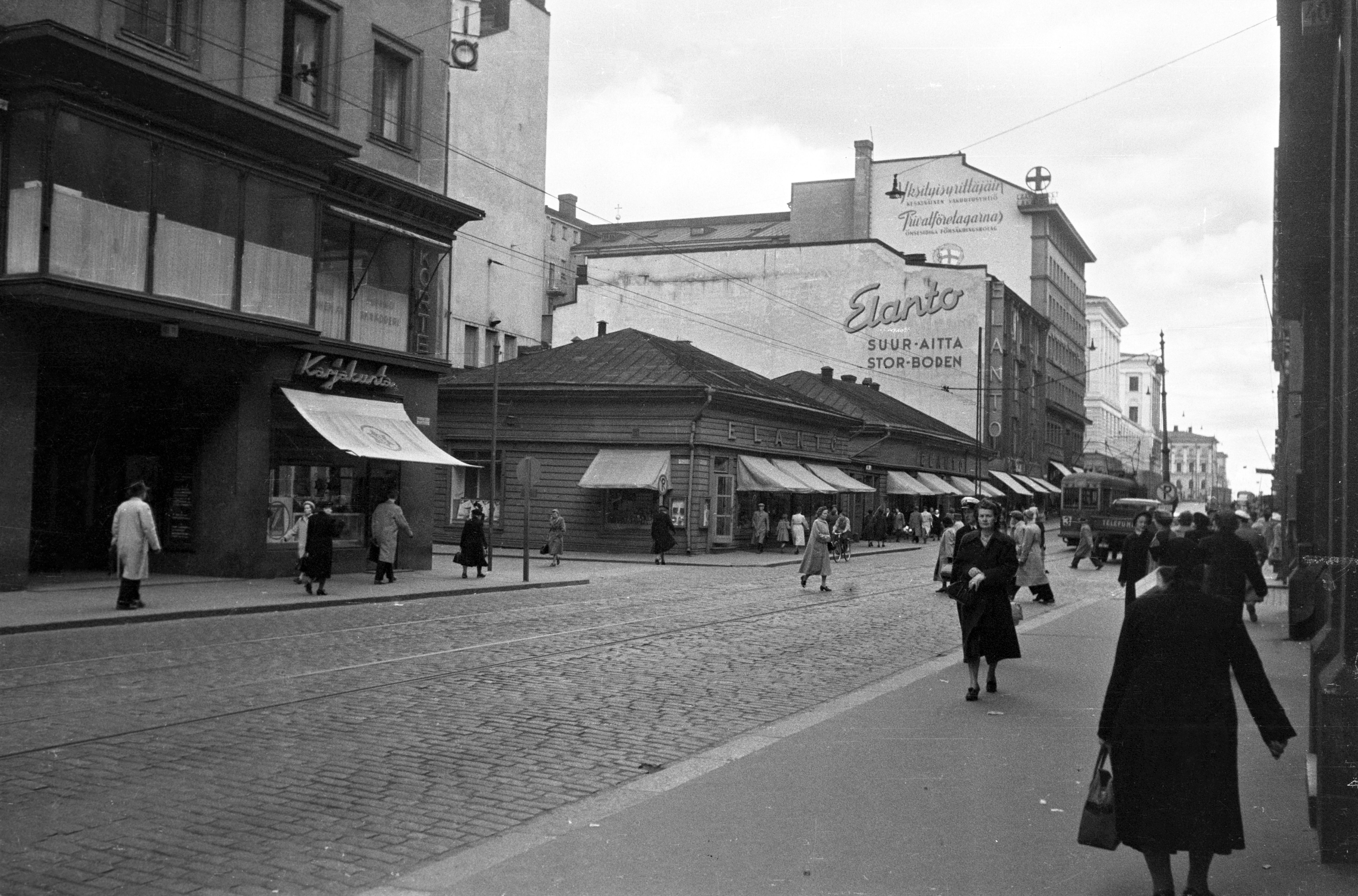 Aleksanterinkatu 11 - 7. - Kluuvikatu 5. Näkymä Aleksanterinkatua itään. Karjakunta -myymälä, Elannon myymälä ja Elannon suur-aitta. Puurakennukset purettiin syksyllä 1950 ja paikalle rakennettiin Elannon tavaratalo.