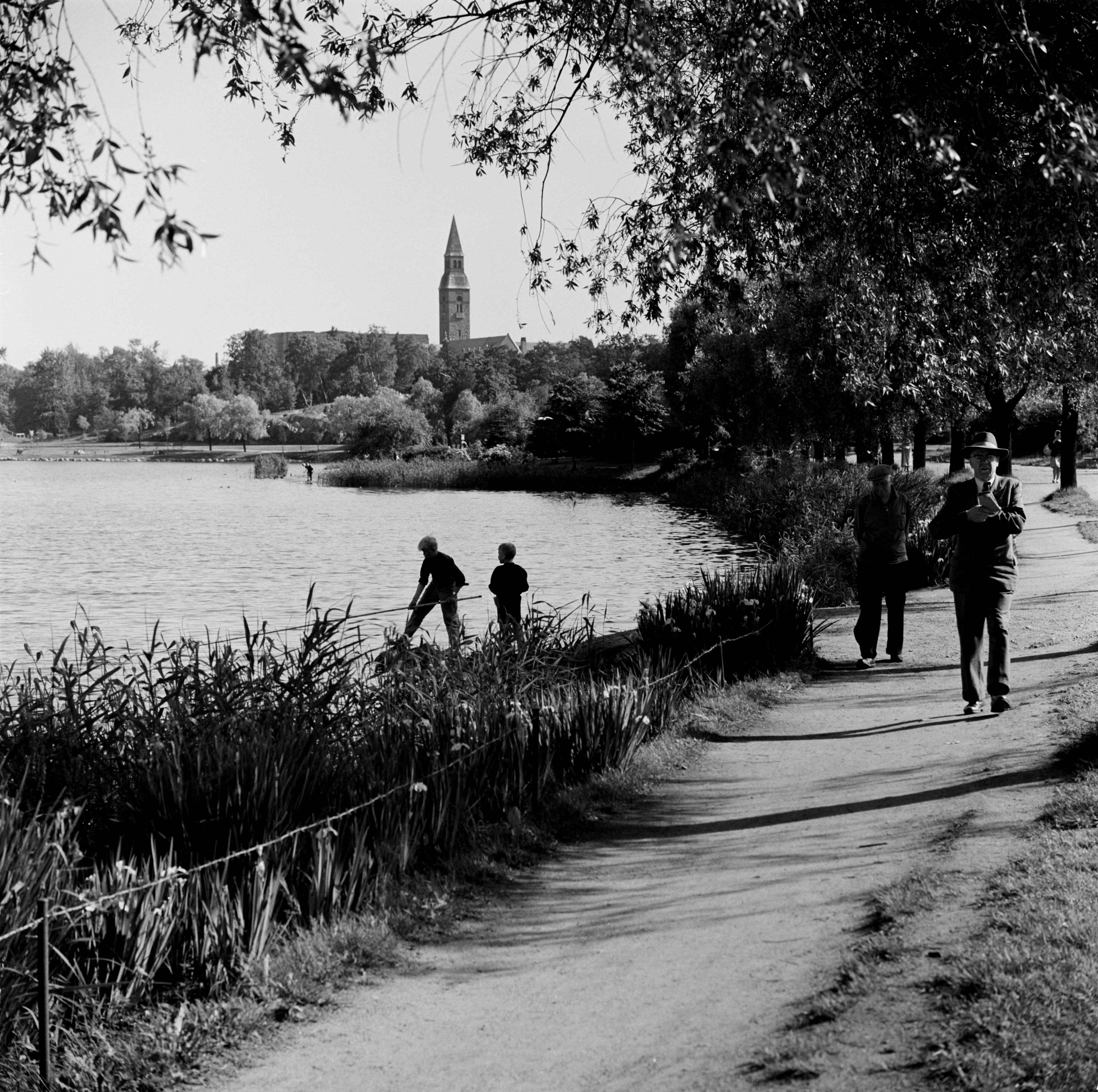 Jalankulkijoita Hesperian puiston rantatiellä ja pojat ongella Töölönlahden rannalla.