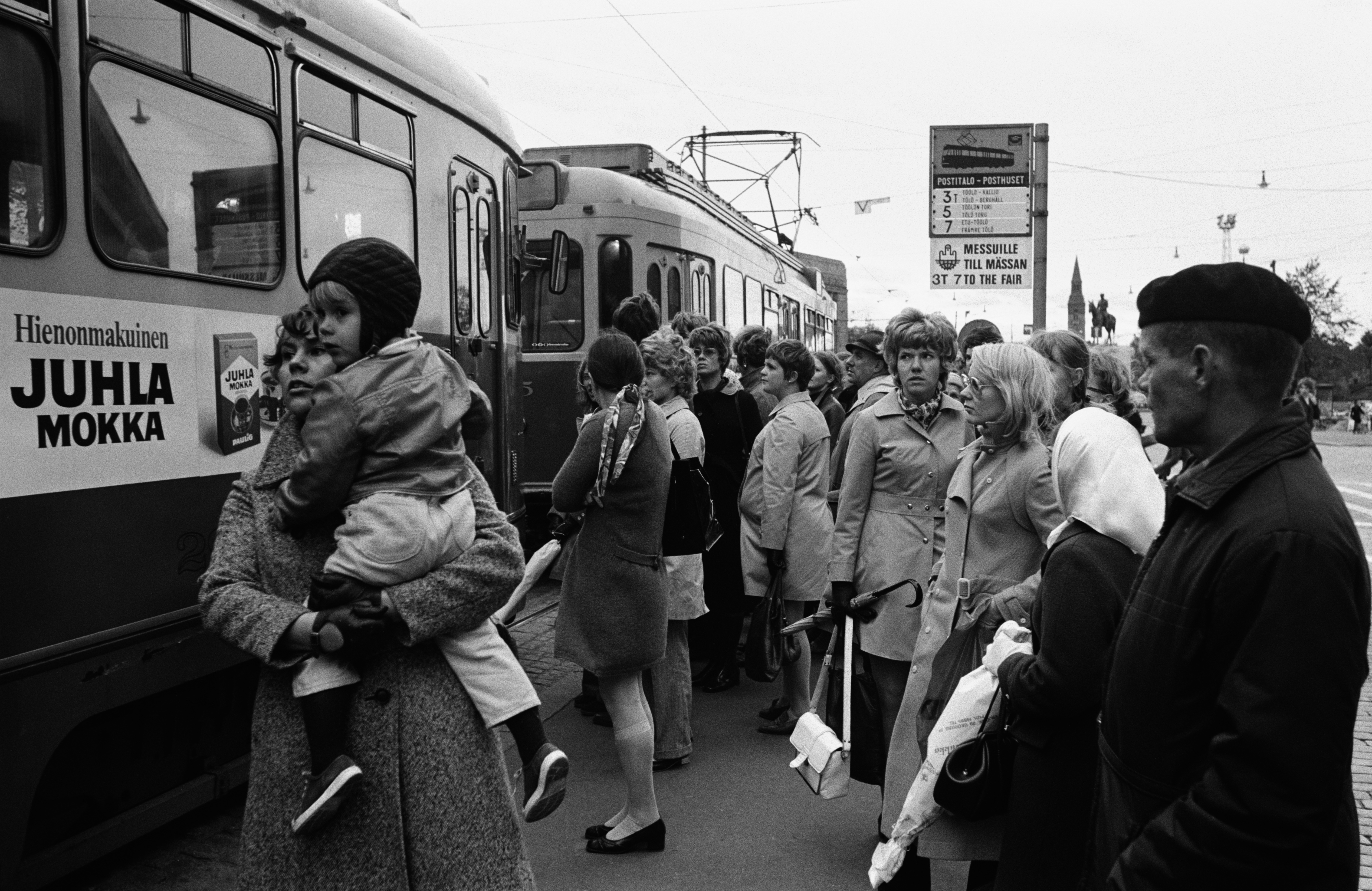 Nainen lapsi sylissään, muita ihmisiä ja kaksi raitiovaunua raitiovaunupysäkillä ruuhka-aikaan Mannerheimintiellä Sokoksen edustalla.