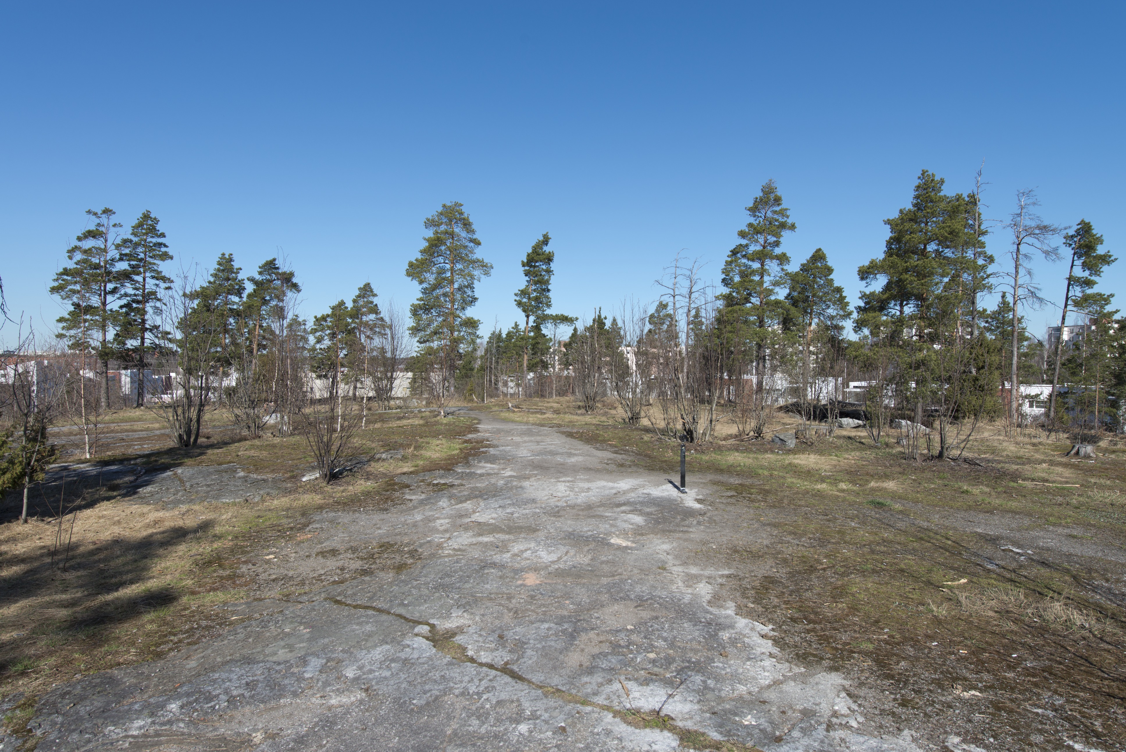 Länsimäki. Ensimmäisen maailmansodan aikainen maalinnoitus, puolustusasema II:8 Länsimäessä, Vantaalla. Kuvassa on aseman ympäristöä. Laakean kallion yli kulkee polku, joka on merkitty metallisin palkein vierailijoiden alueella suunnistusta helpottamaan. Kuvan keskellä näkyy yksi näistä palkeista. Taustalla on asuinrakennuksia.