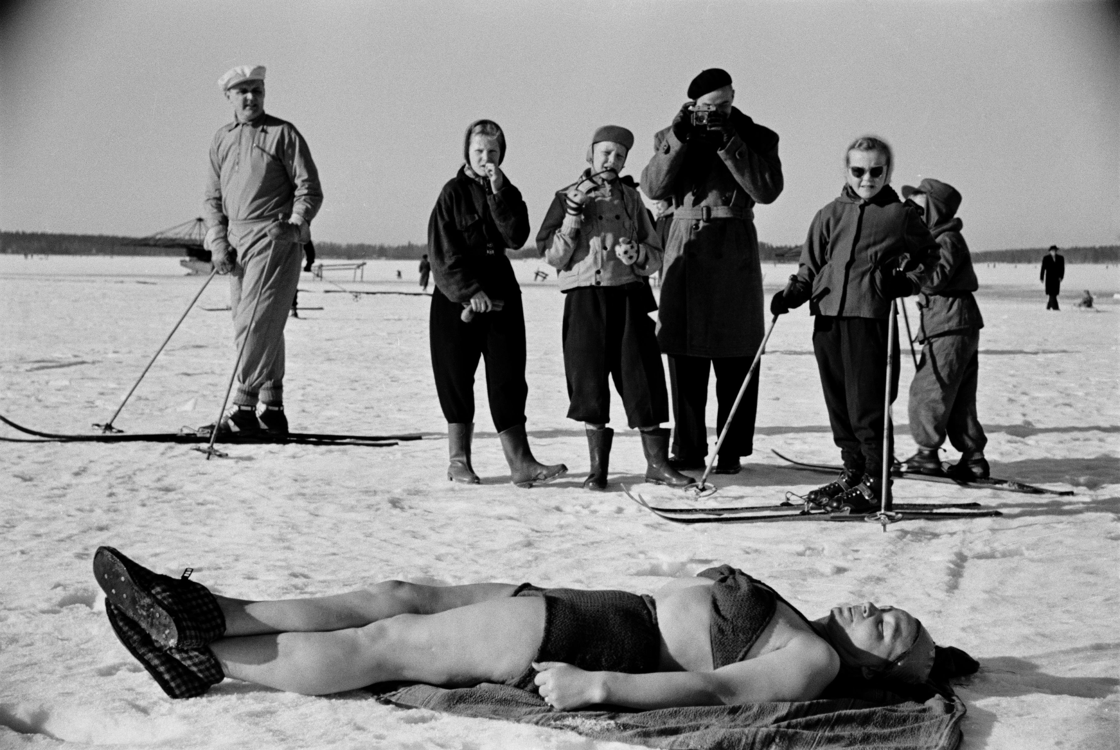Avantouintia Taivallahdella. Uimarit ottavat aurinkoa uinnin jälkeen, nainen virkatuissa bikineissä. Ympärillä yleisöä, mm. hiihtäjiä ja valokuvaa ottava mies.