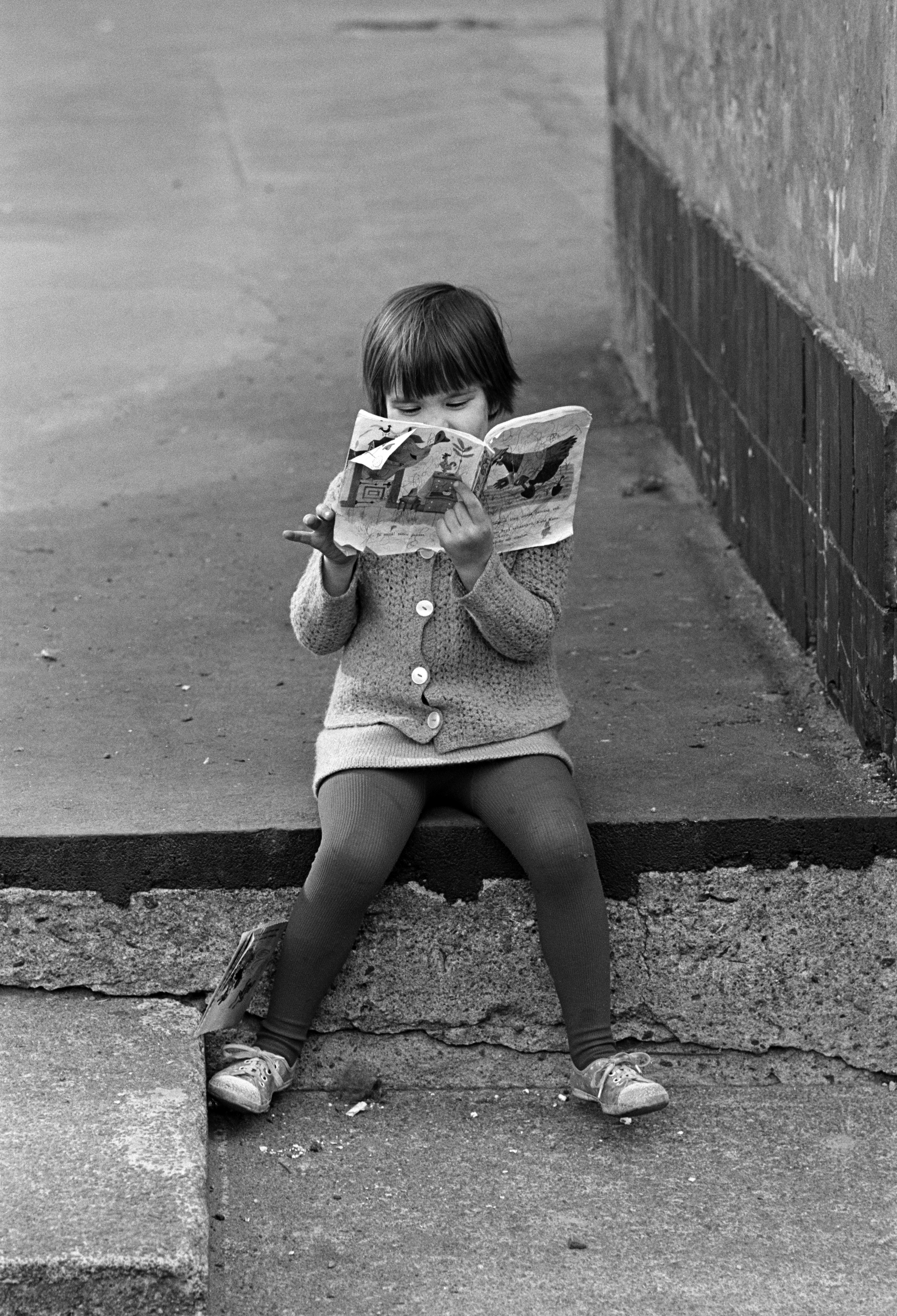 Repaleista kirjaa lukeva tyttö istumassa Viides linja 4:n pihalla. Tytöllä yllään neuletakki.