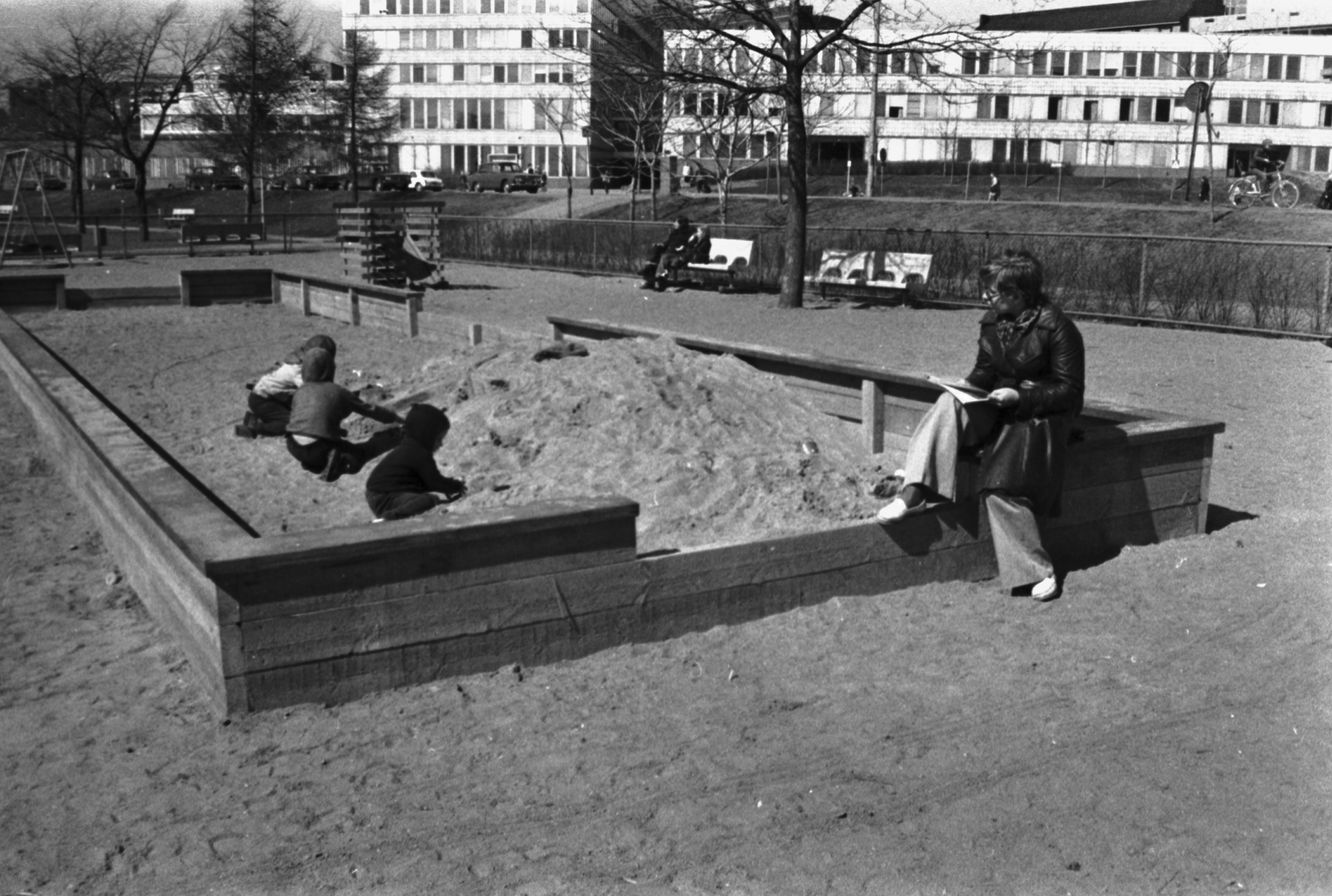 Tokoinrannan puisto. Nainen ja lapsia leikkimässä suuressa hiekkalaatikossa Eläintarhantien ja Säästöpankinrannan kulmassa olevassa puistossa. Hiekkalaatikon reunalla istuu lehteä lukeva nainen. Taustalla Kallion virastotalo.