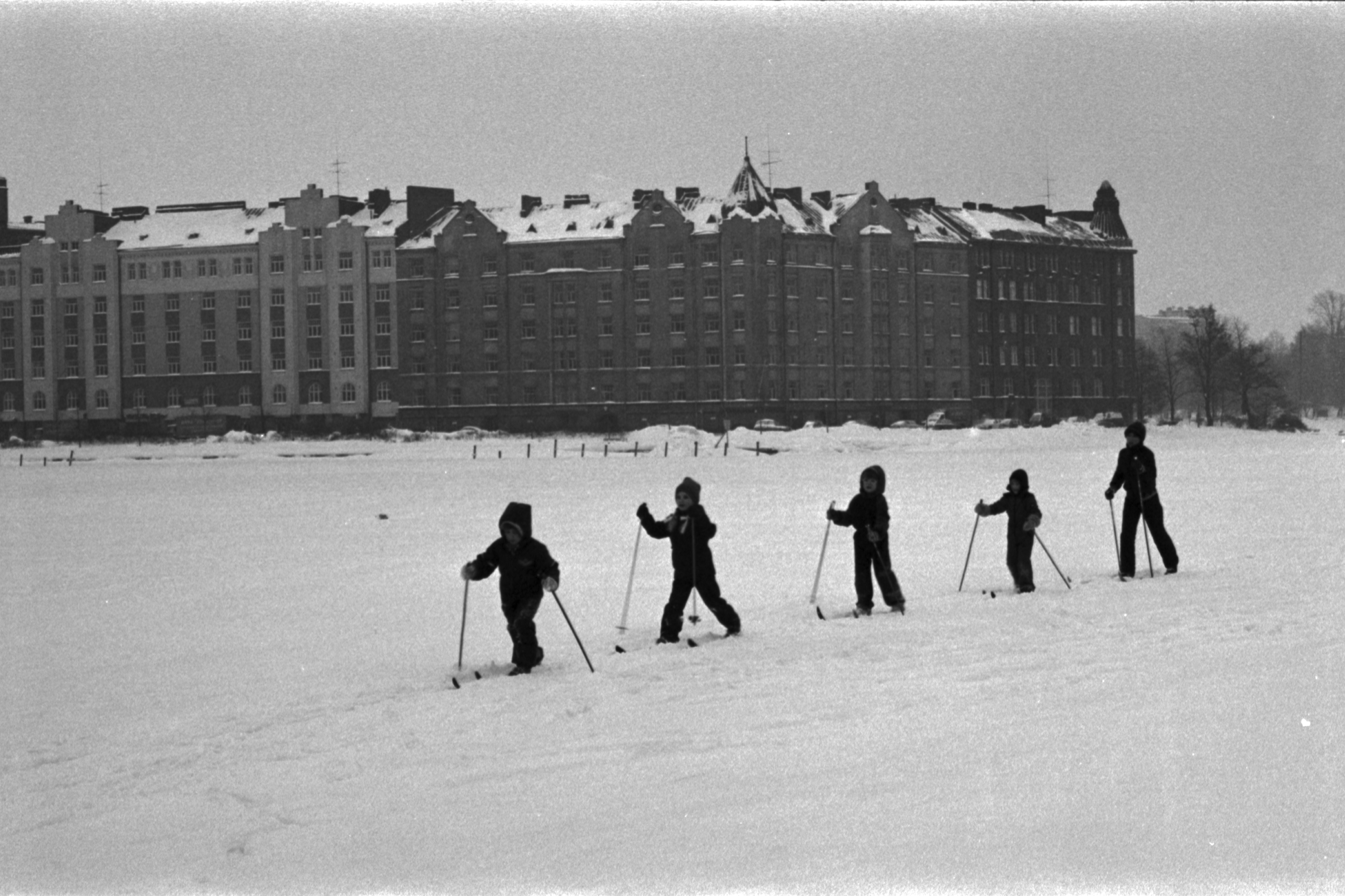 Eläintarhanlahti. Vironniemen päiväkodin lapsia, jonon päässä opettaja, hiihtämässä Eläintarhanlahden lumisella jäällä sumuisella säällä kohti Tokoinrantaa. Taustalla Siltasaari.