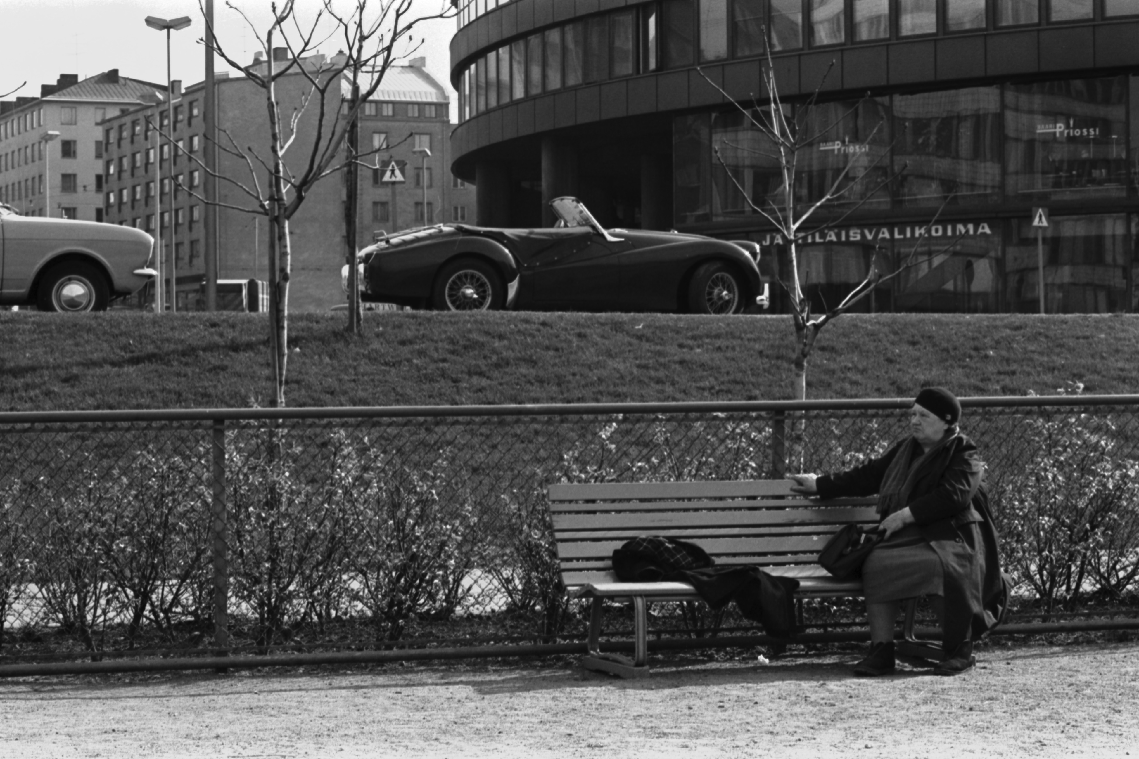 Tokoinranta. Eläintarhantie. Vanhahko nainen istumassa penkillä Tokoinrannan puistossa. Taustalla pysäköity urheiluauto (Triumph TR3) Eläintarhantiellä Ympyrätalon edessä. Vasemmassa reunassa näkyy Ford Cortina.