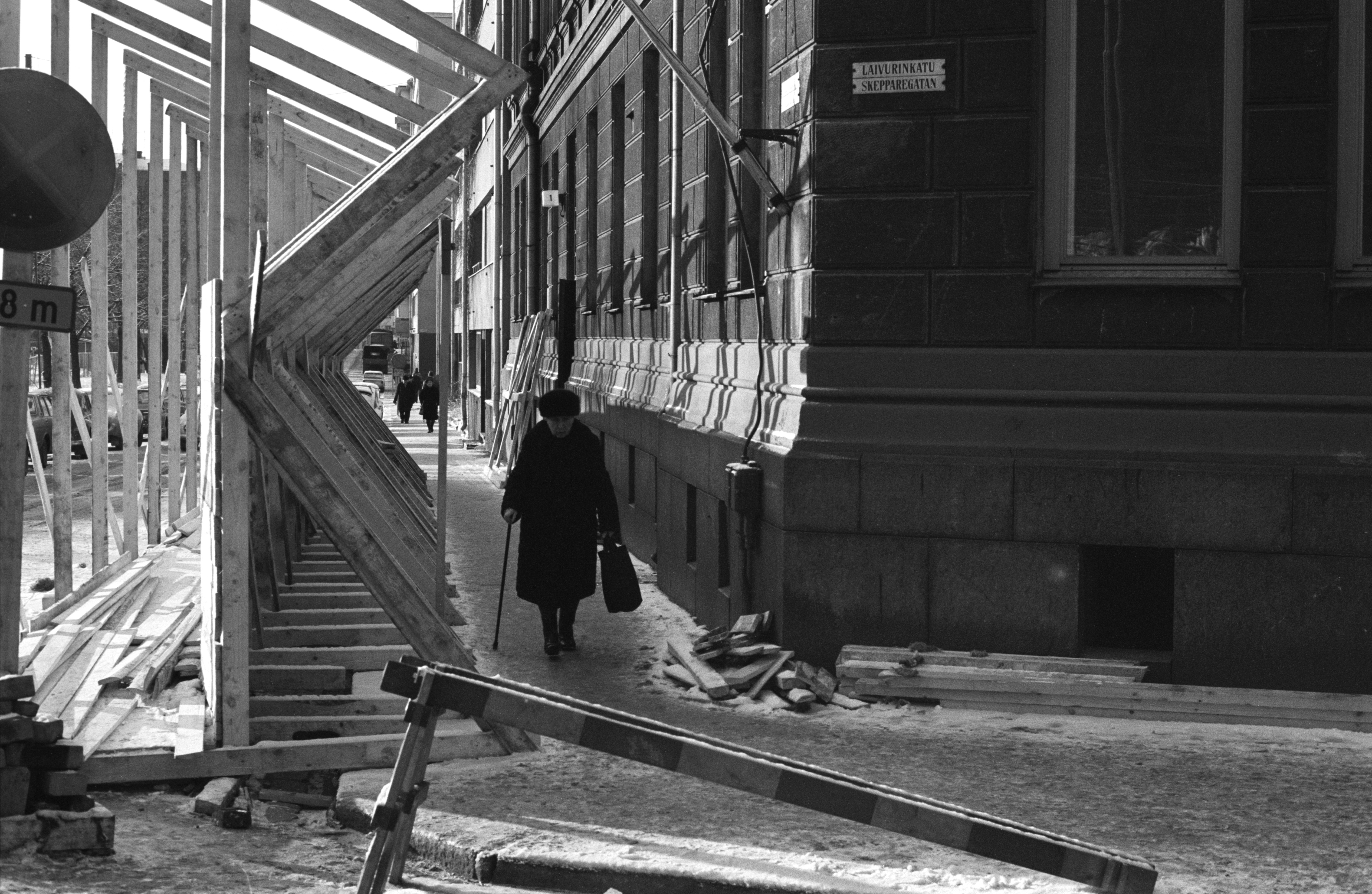 Vanha nainen kepin kanssa kävelemässä purettavan kerrostalon edustalle rakennetulla puisella jalankulkukäytävällä Laivurinkadun ja Sepänkadun kulmassa.