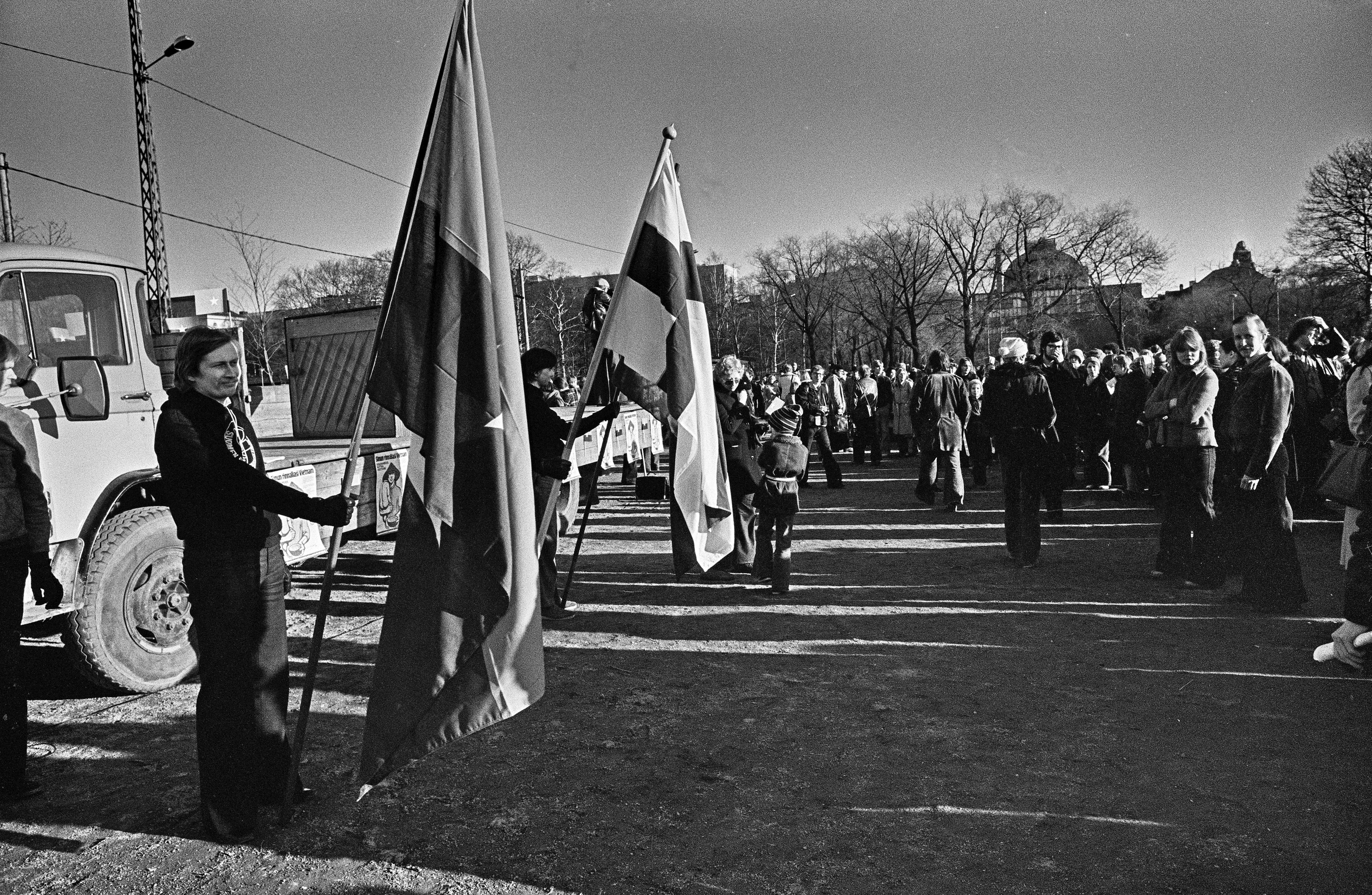Mielenosoittajia kokoontuneena Kaisaniemen urheilukentälle. Yksi mielenosoittaja pitää pystyssä tangossa olevaa Suomen lippua ja toinen Etelä-Vietnamin kansallisen vapautusrintaman lippua.