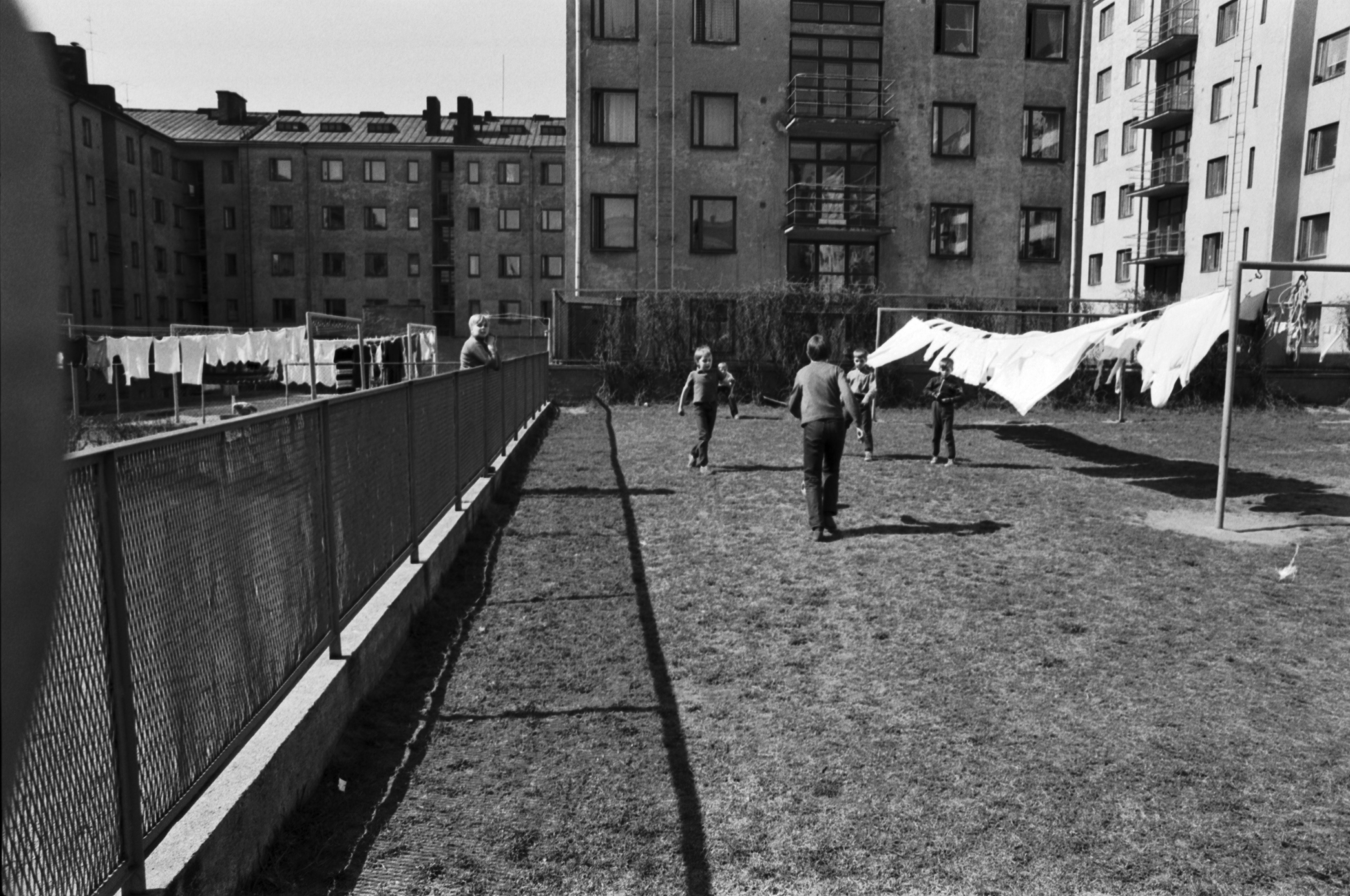 Neljäs linja 24. Näkymä Kolmas linja - Castréninkatu - Neljäs linja - Suonionkatu -katujen ympäröimän korttelin pihalta luoteeseen. Etualalla poikia pelaamassa jalkapalloa Neljäs linja 24:n nurmikkopihalla valkopyykin liehuessa narulla kuivumassa. Taaempana Neljäs linja 26 talo. Vasemmalla verkkoaidan takana Kolmas linja 25 pyykkejä. Taustalla Castréninkatu 3.