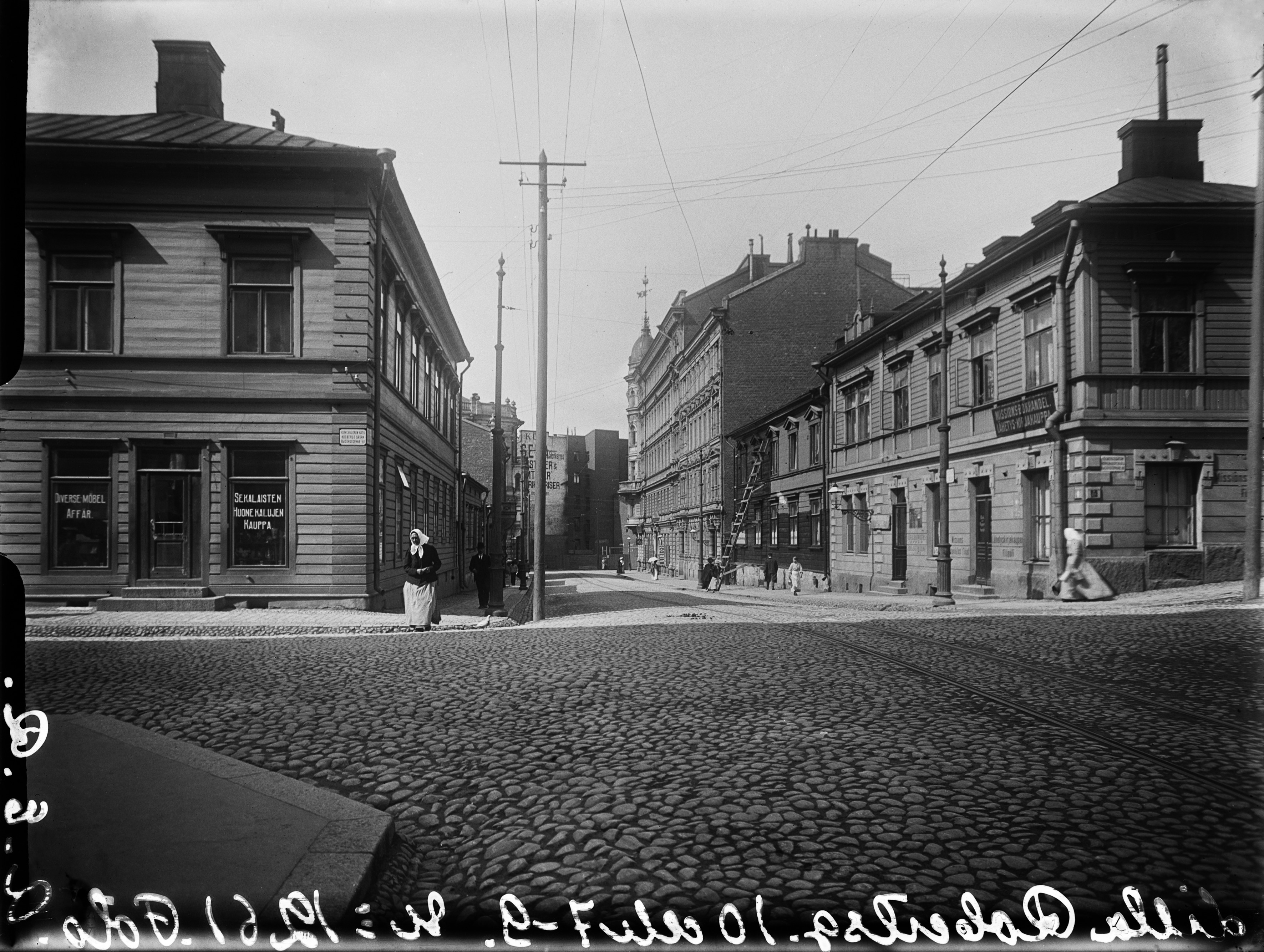 Vasemmalla Pieni Roobertinkatu 10 (- Korkeavuorenkatu 20), oikealla Pieni Roobertinkatu 7 (- Korkeavuorenkatu 22), 9, 11, 13. Liikehuoneistoissa mm. Sekalaisten Huonekalujen Kauppa ja Lähetys-Kirjakauppa, kadulla jalankulkijoita.
