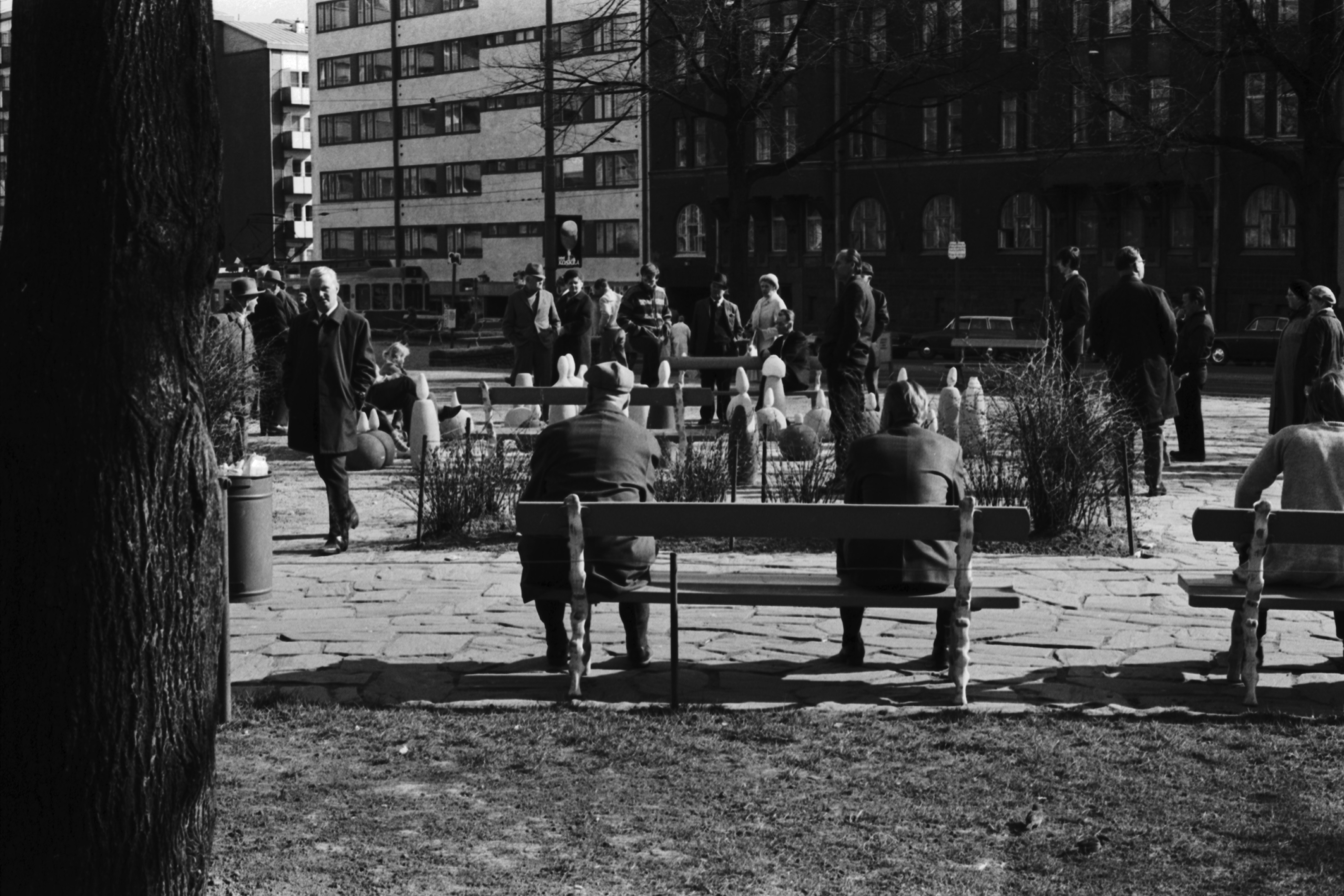 Karhupuisto. Ihmisiä istuskelemassa penkeillä sekä pelaamassa shakkia aurinkoisessa Karhupuistossa.