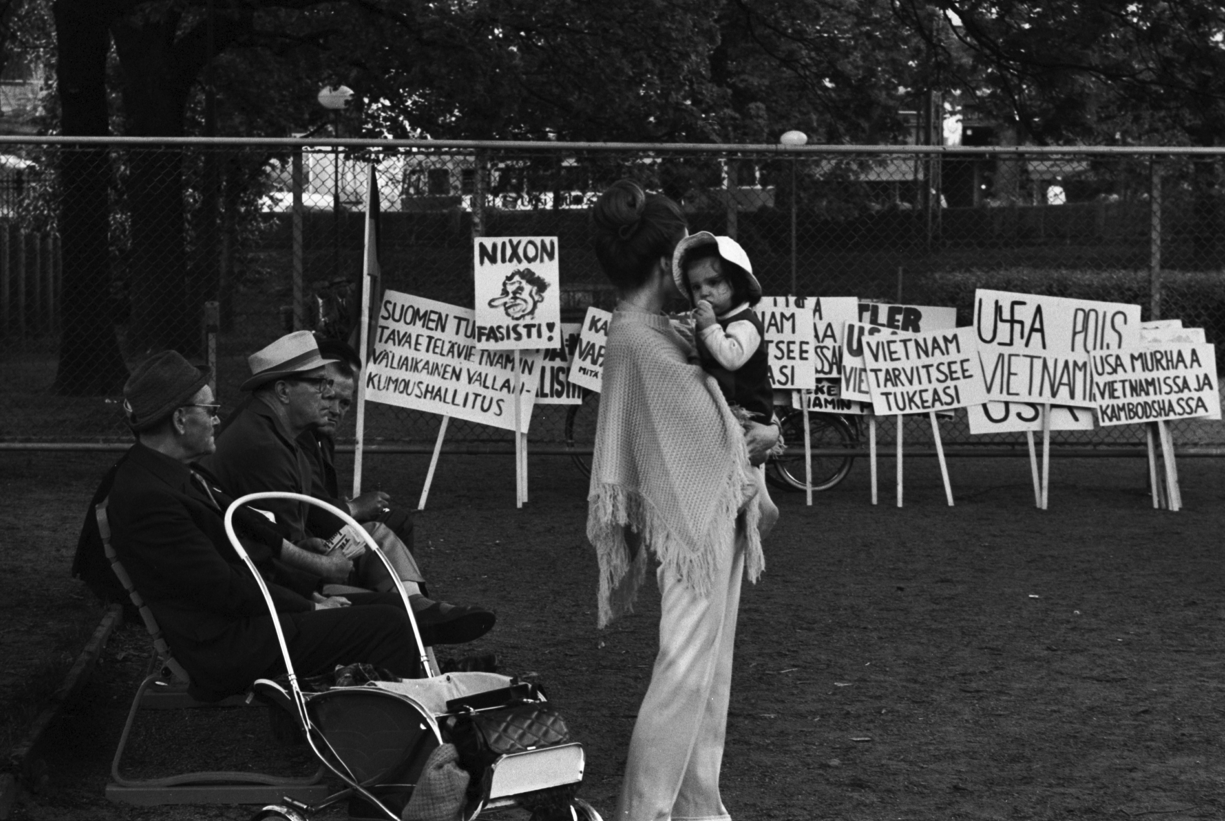 Kaisaniemenpuisto. Nainen lapsi sylissä ja penkillä istuvia miehiä sekä aitaa vasten nojallaan olevia Vietnamin sodan vastaisia mielenosoituskylttejä Kaisaniemenpuistossa. Puistossa meneillään Vietnamin sodan ja imperialismin vastainen mielenosoitus.