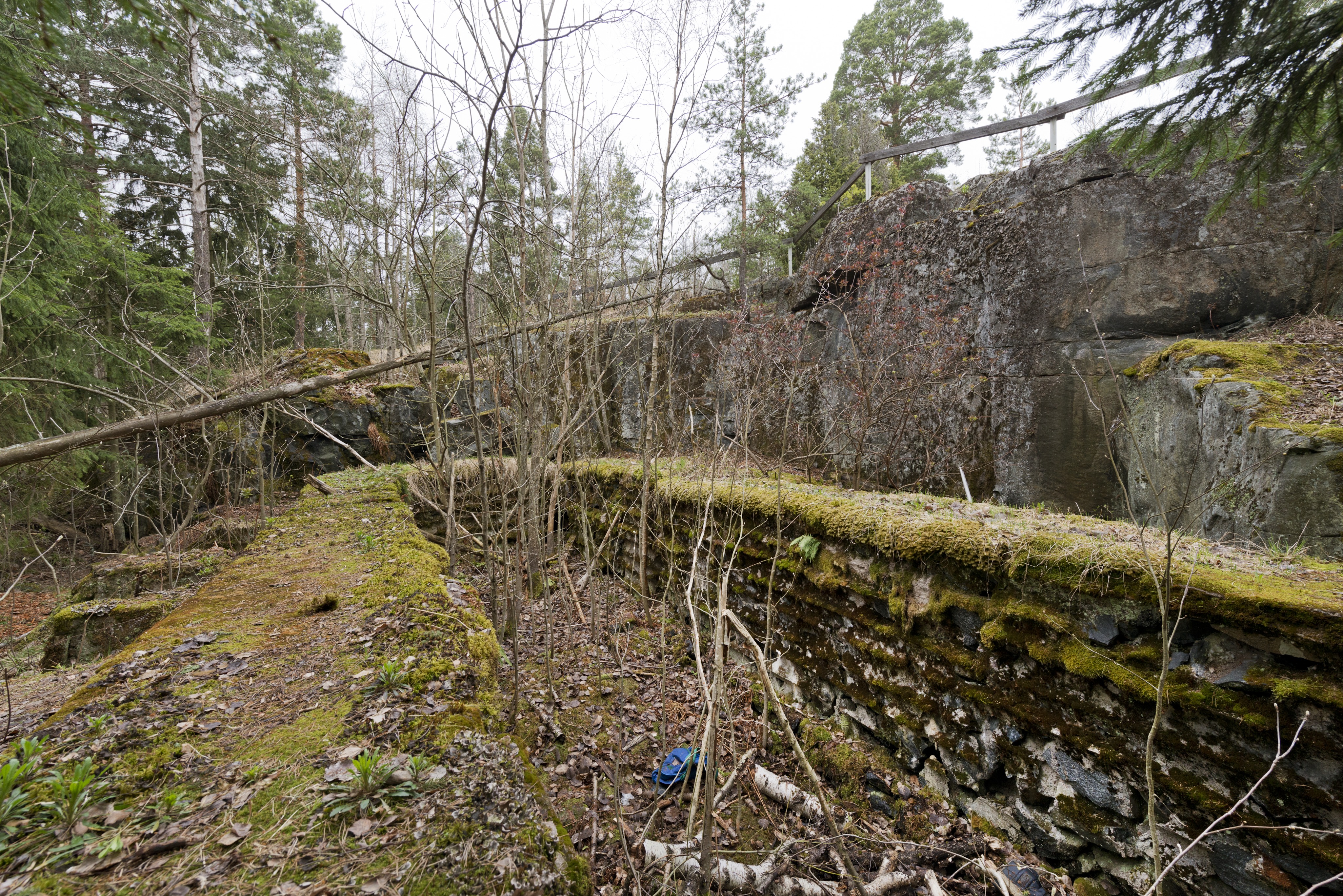 Mäkkylä. Ensimmäisen maailmansodan aikainen maalinnoitus, puolustusasema XXVIII:19 Mäkkylässä, Espoossa. Kuvassa on aseman lounaisosassa sijaitseva tuliasemarakenne neljälle kiväärimiehelle ja yhdelle konekiväärille, kuvassa näkyvät vasemmalla ampuma-aukot. Rakenne on tehty valamalla betonia hirsikehikoiden keskelle. Asema oli myös katettu hirrellä, joka peitettiin maalla. Kaikki hirsirakenteet on myöhemmin purettu ja maa-ainekset kasattu ampuma-aukkojen eteen. Seinissä erottuu hirsirakenteista jääneet painaumat. Aseman takana olevan kallion reunaa kiertää puinen kaide. Aseman pohjalla kasvaa vesakkoa. Kuvaussuunta länteen.