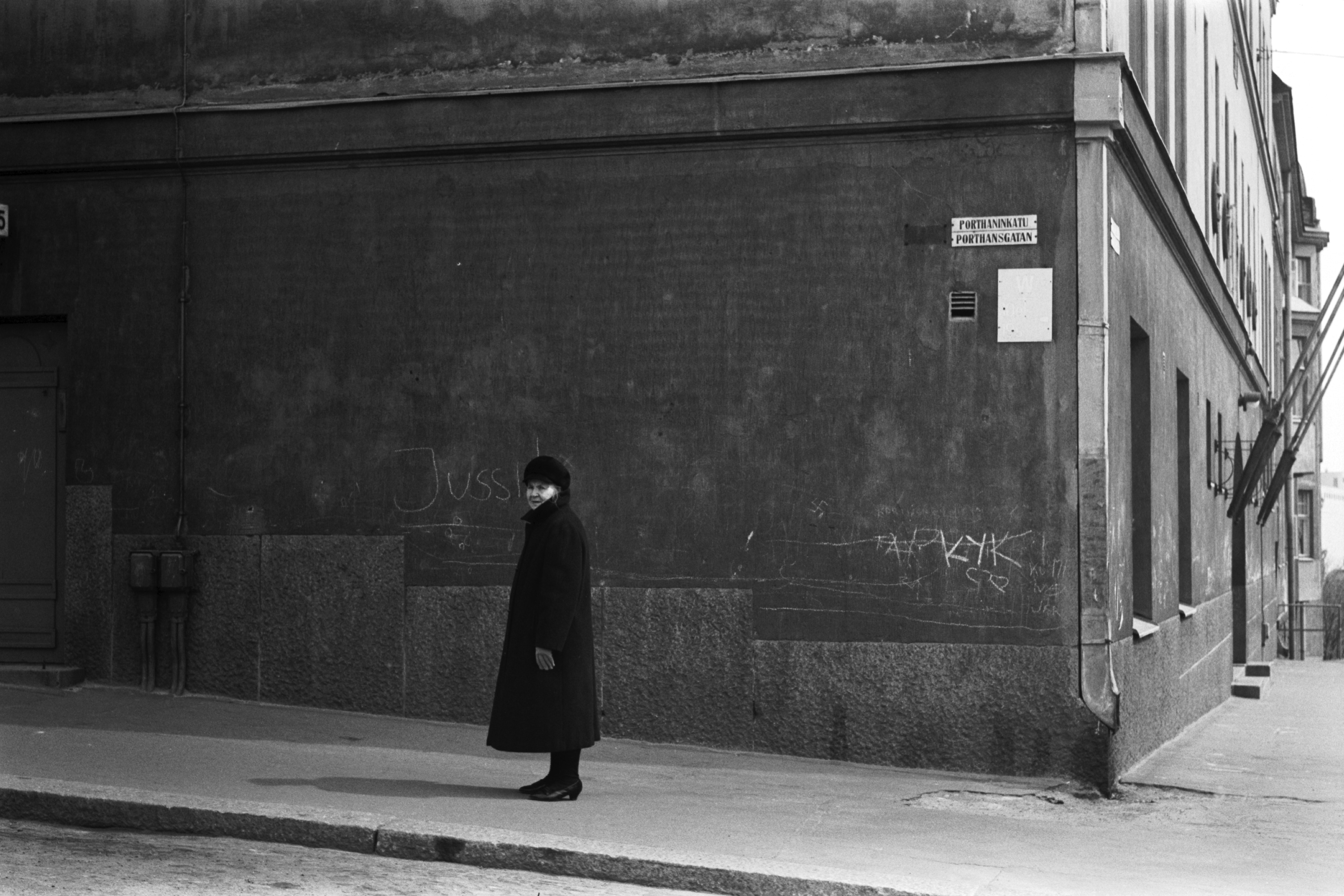 Porthaninkatu 15. Vanha nainen seisomassa jalkakäytävällä Porthaninkadulla Kaikukujan kulmassa.