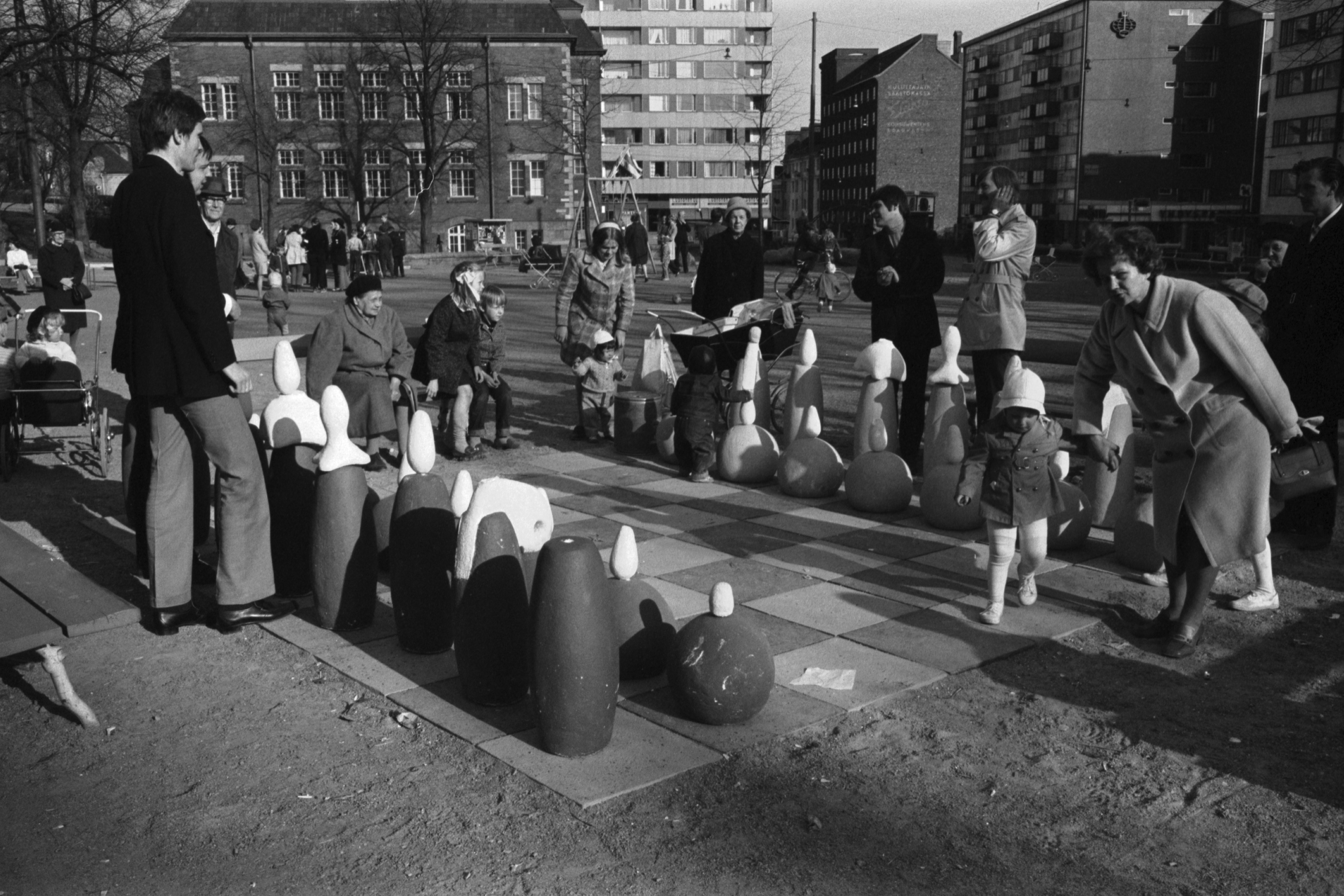 Karhupuisto. Helsingin juhlaviikot. Aikuisia ja lapsia järjestelemässä nappuloita suurella shakkilaudalla Karhupuistossa.