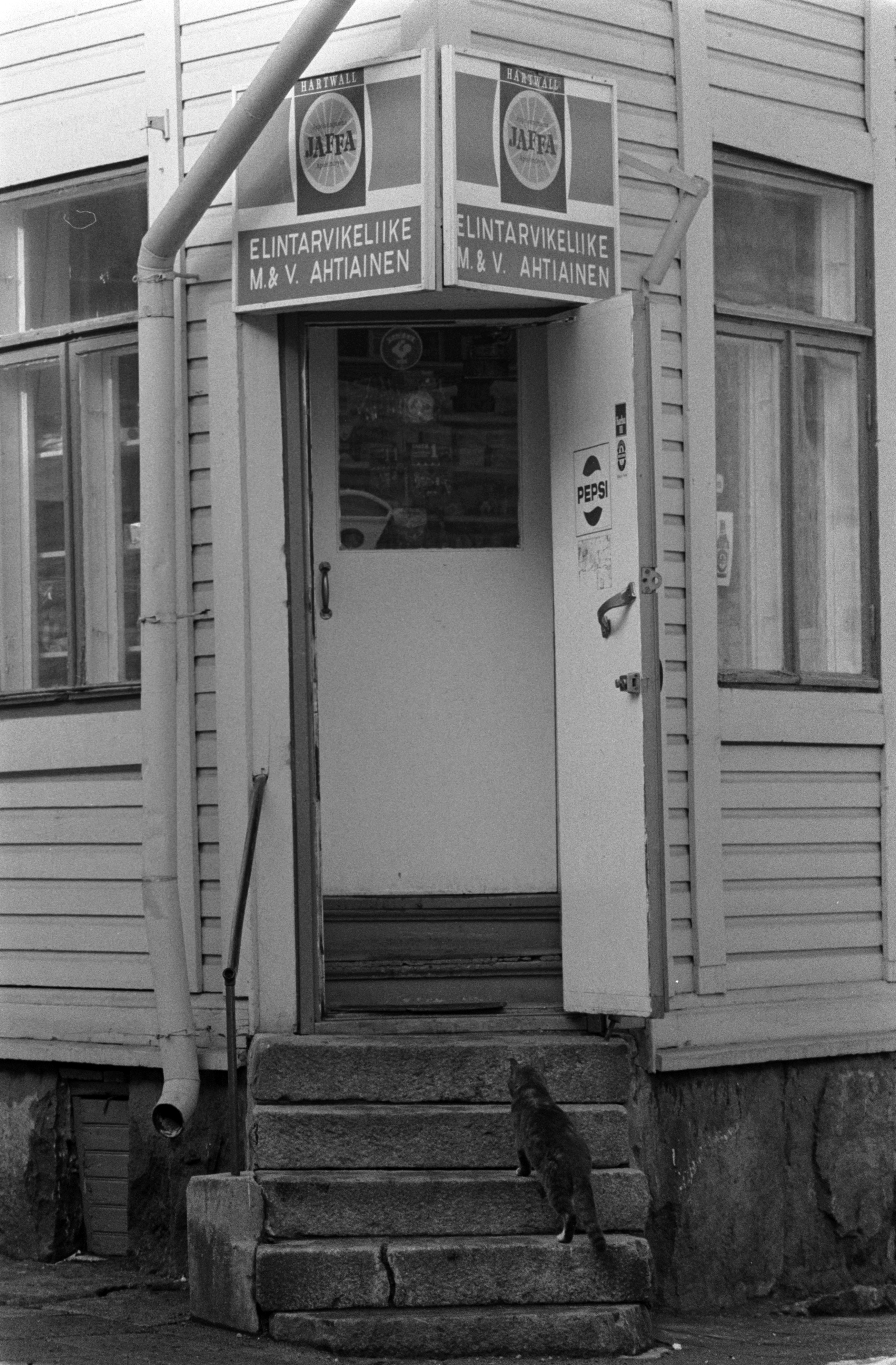 Elintarvikeliike M & V. Ahtiainen Vallilantien ja Keuruuntien kulmassa sijaitsevassa puutalossa Puu-Vallilassa. Kissa kiipeää portaita pitkin kohti liikkeen avointa ovea.