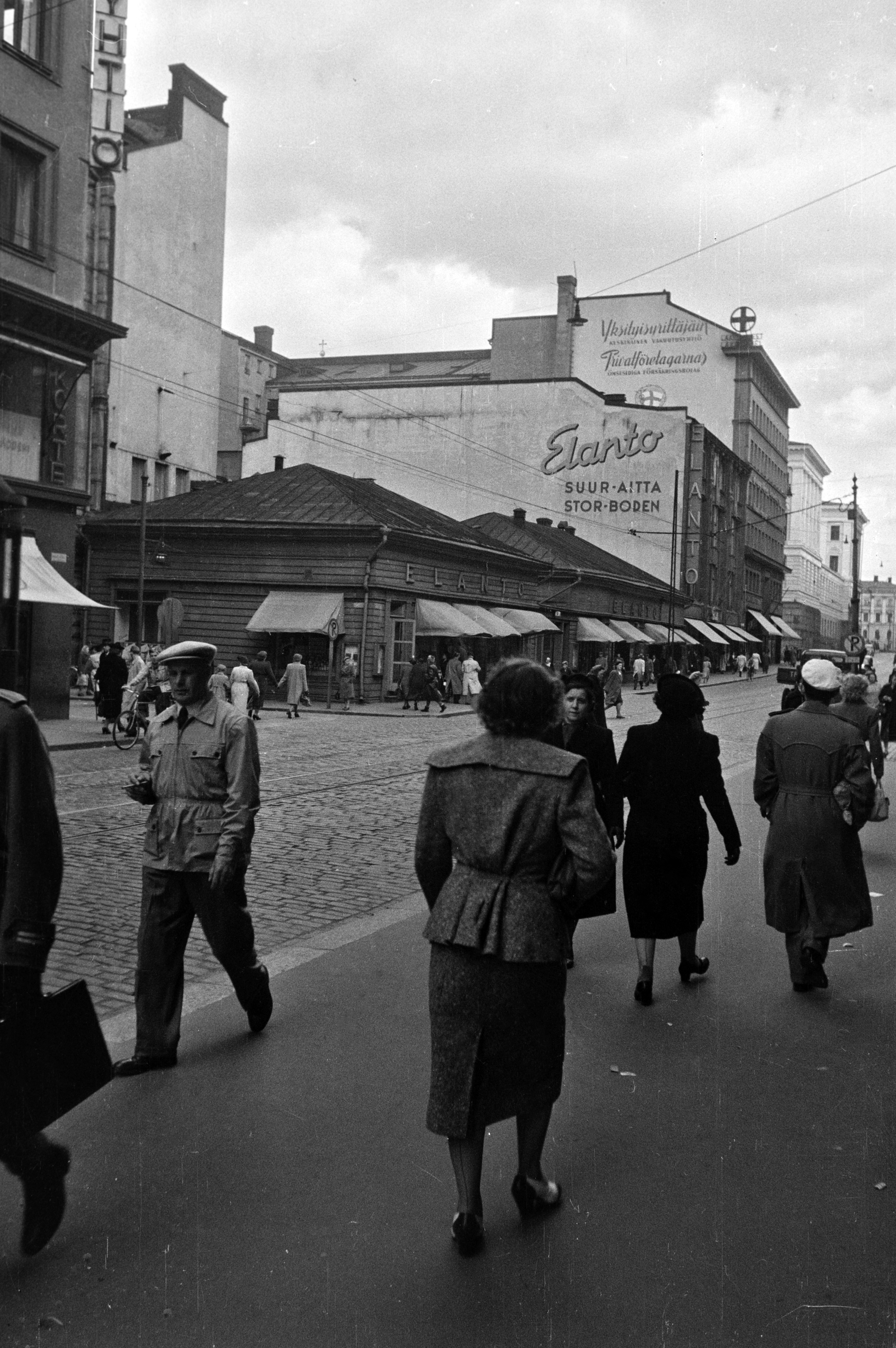 Kluuvikatu 5. - Aleksanterinkatu 9, 7. Näkymä Aleksanterinkatua itään. Elannon myymälä ja Elannon suur-aitta. Puurakennukset purettiin syksyllä 1950 ja paikalle rakennettiin Elannon tavaratalo.
