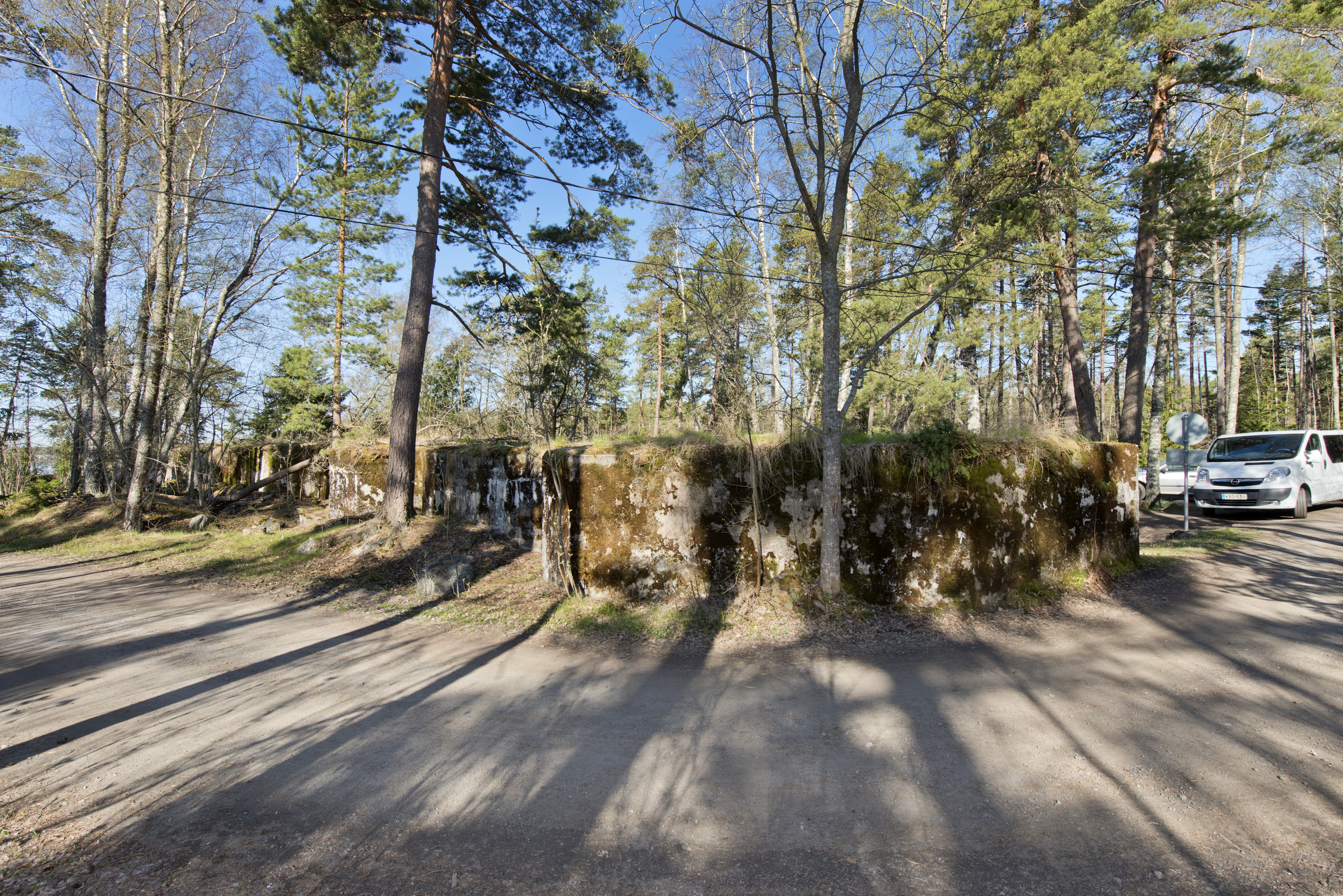 Skatanniemi. Ensimmäisen maailmansodan aikainen rannikkotykkipatteri Skatanniemessä, Vuosaaressa. Kuvassa on rintasuoja, jonka taakse on sijoitettu yhteensä neljä tykkiasemaa. Rintasuoja aaltoilee loivasti asemien kohdalta. Seinät ovat sammaleen peitossa. Kuvan etualalla kulkee tie, joka on puhkaistu aseman läpi 1920-luvulla. Taustalla oikealla näkyy autoja. Kuvaussuunta suurin piirtein länteen.