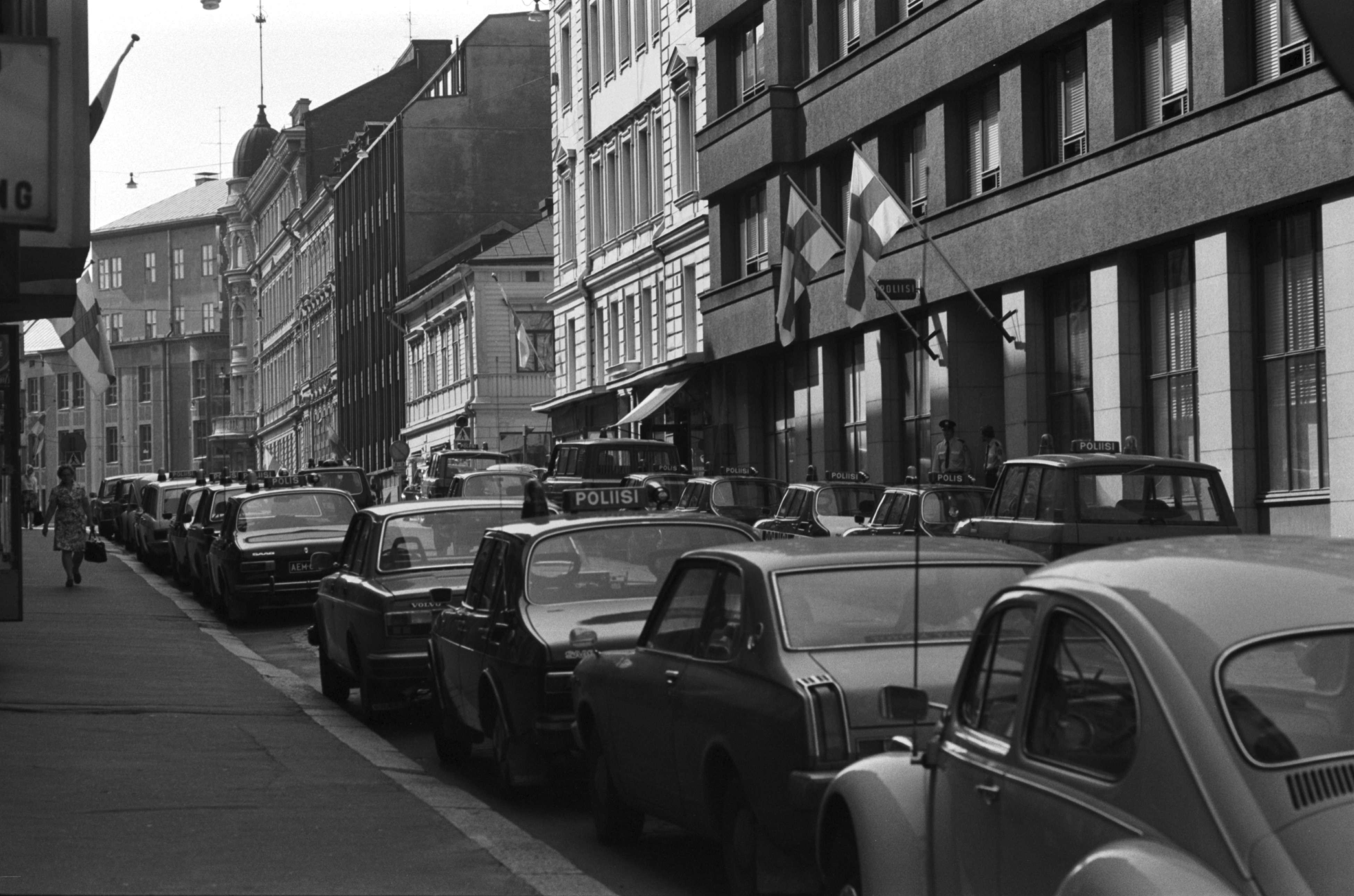 Pieni Roobertinkatu 1-3. Näkymä Pieneltä Roobertinkadulta Kasarmikadun kulmasta länteen Korkeavuorenkadun suuntaan. Liputetun poliisiaseman edustalla kaksi poliisia ja poliisiautoja. Euroopan turvallisuus- ja yhteistyökokous (ETYK) järjestettiin Helsingissä Finlandia-talossa heinä-elokuun vaihteessa 1975.