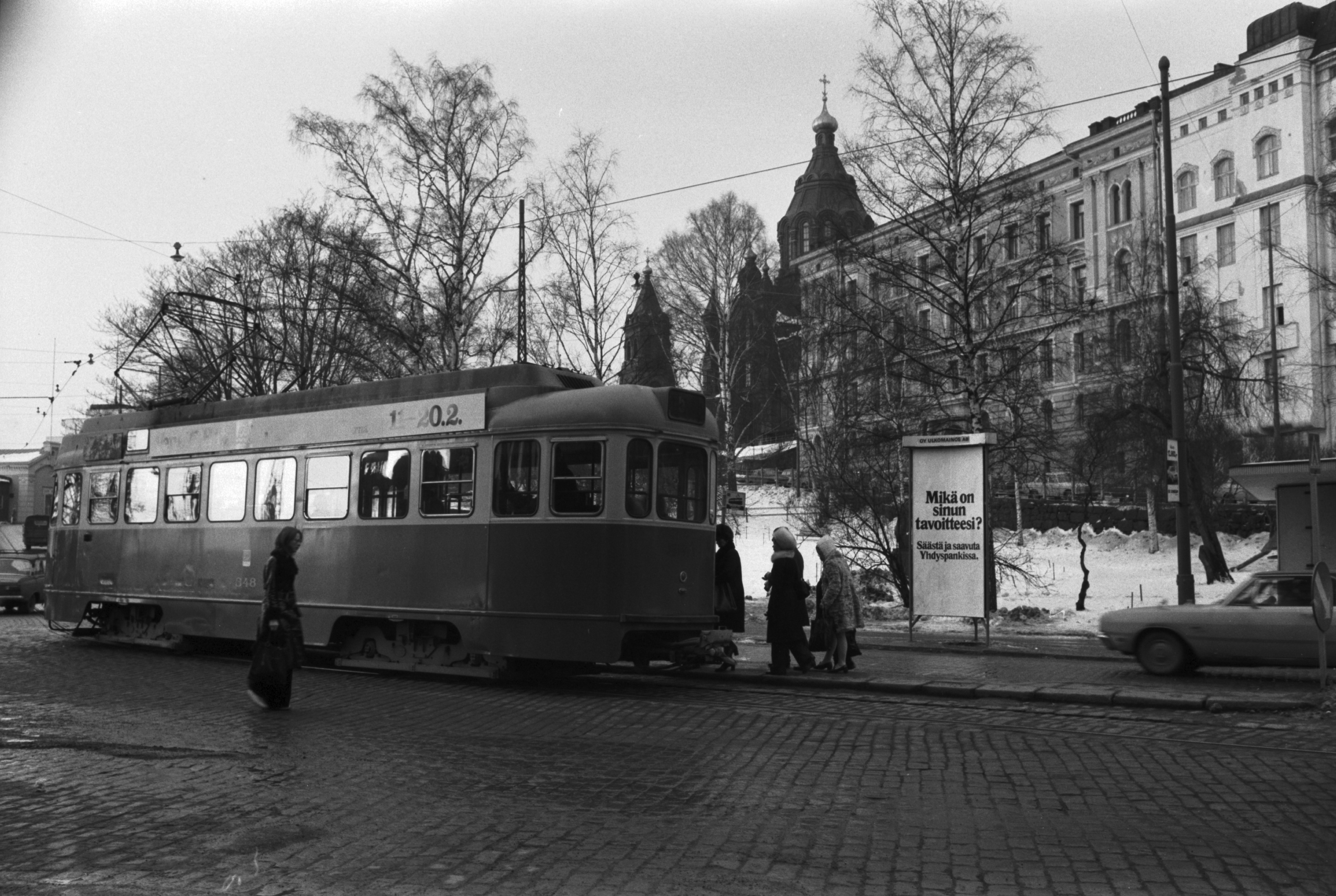 Katajanokka, Satamakatu. Matkustajia kapealla pysäkkikorokkeella nousemassa linjan 5 raitiovaunuun numero 348 pysäkillä Satamakadulla Katajanokan puiston (nykyisin Tove Janssonin puisto) laidalla. Taustalla Rahapajankatu sekä Uspenskin katedraali. Kolmikulmaisessa ulkomainostelineessä Pohjoismaisen Yhdyspankin mainos.