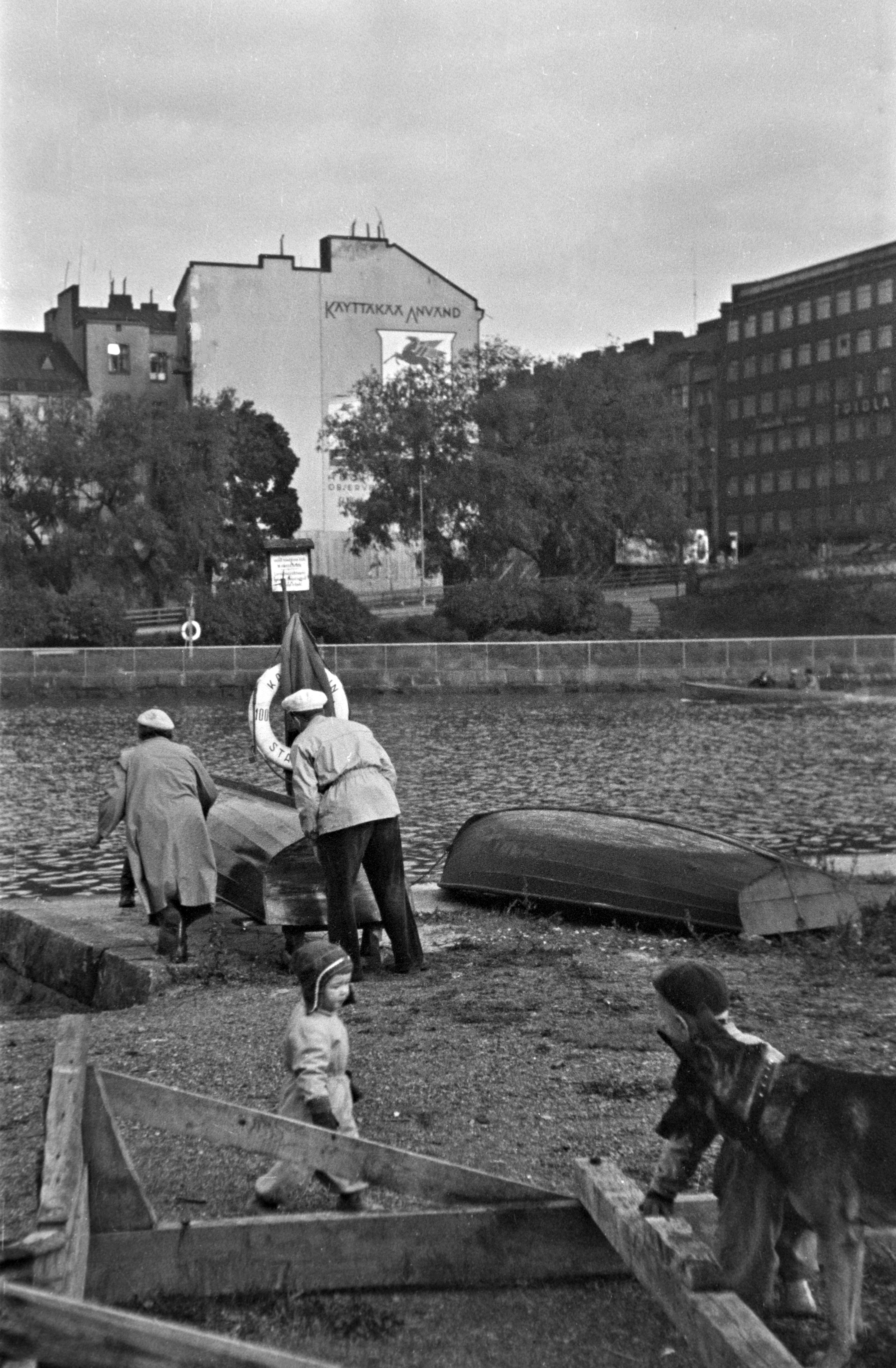 Kaksi henkilöä nostamassa venettä Kaisaniemenlahden rannalla. Etualalla kaksi lasta ja koira. Taustalla Pitkänsillanranta 3 ja Siltasaarenkatu 1.