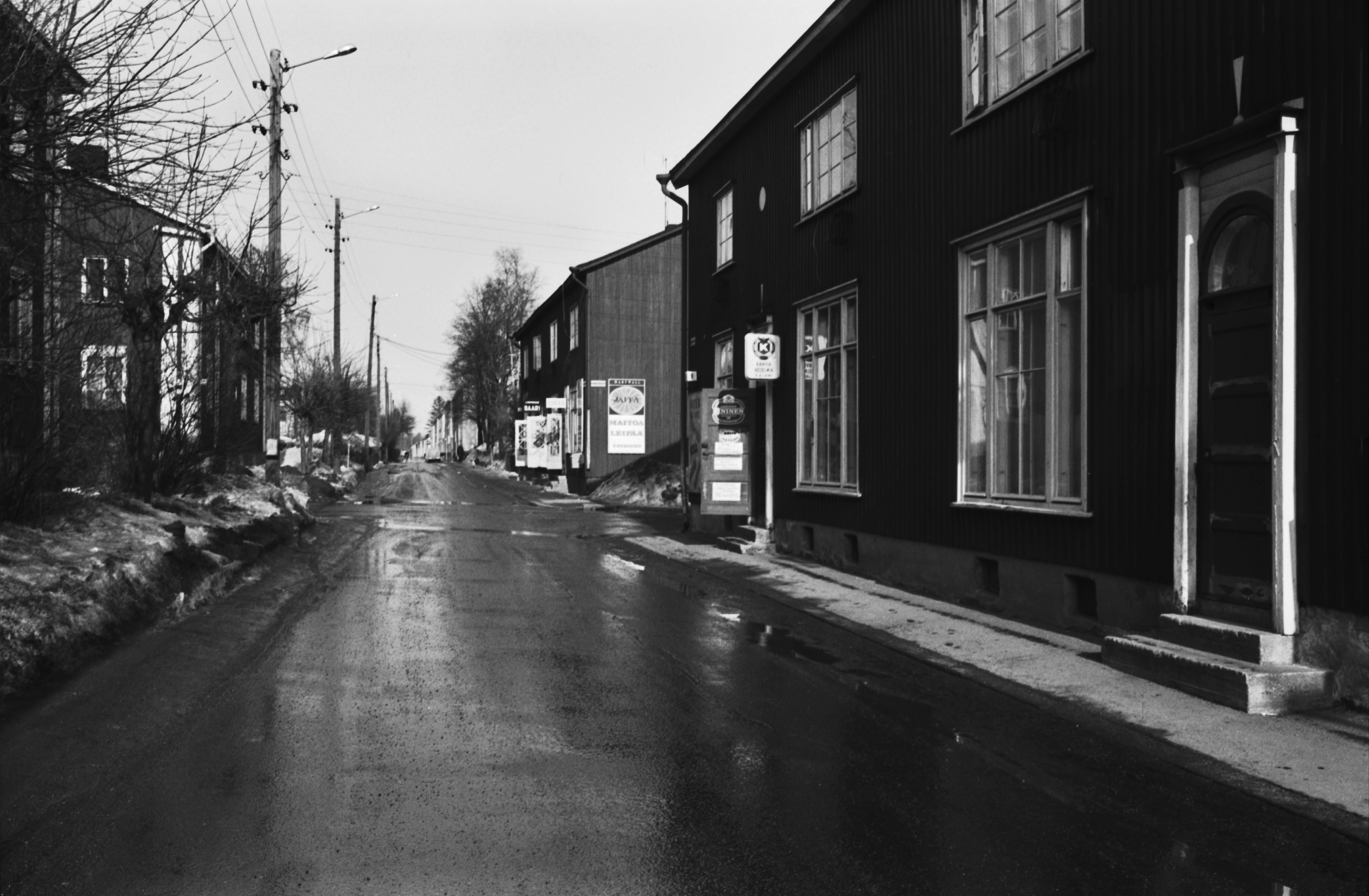 Limingantie 9. Katunäkymä Limingantieltä Jyrängöntien kulmasta koilliseen kevätauringon sulattaessa lumia. Oikealla olevissa kaksikerroksisissa puutaloissa myymälöitä, edessä Limingantie 9:ssä H.M. Laineen K-kauppa.