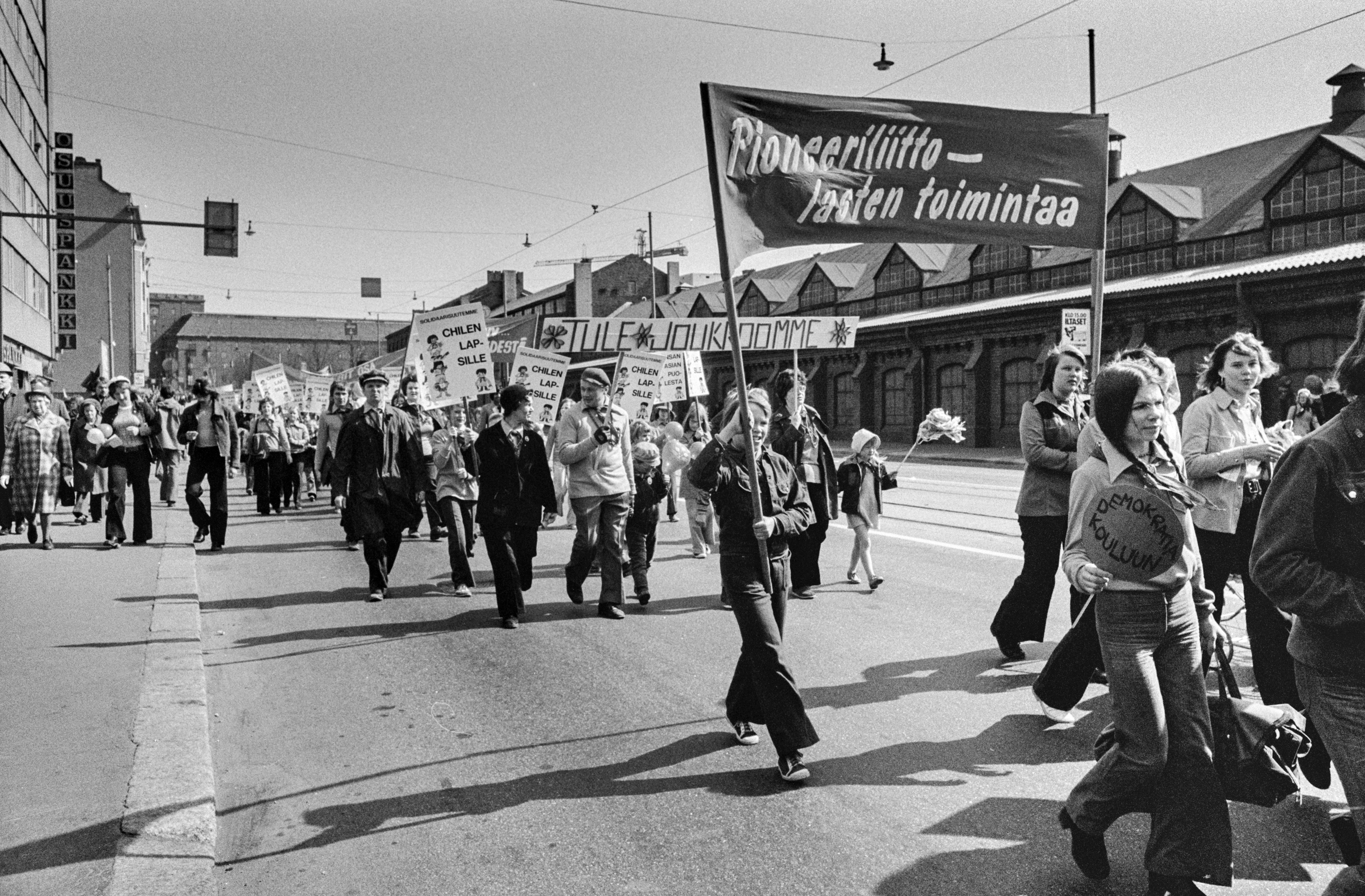 Vappumarssijoita Hämeentiellä. Edessä nuoret kantavat banderollia, jossa teksti: Pioneerilliitto - lasten toimintaa. Takana banderolli, jossa teksti: Tule joukkoomme ja kylttejä, joissa teksti: Solidaarisuutemme Chilen lapsille.