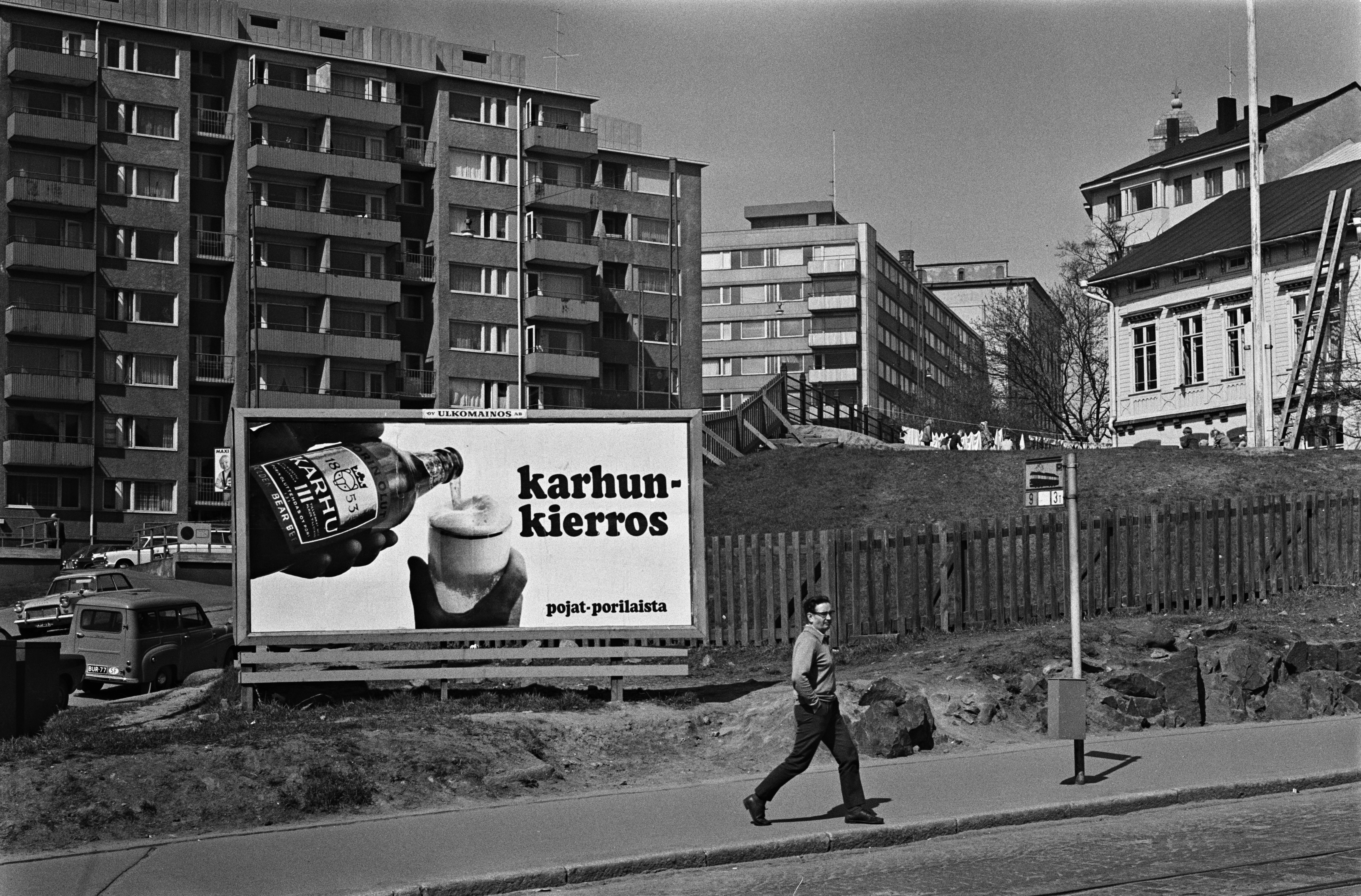 Porthaninkatu 2. Mies kävelemässä raitiovaunupysäkin kohdalla Porthaninkadulla. Tukholman päiväkodin aidan edessä suuri Porin Oluttehtaan Karhu-olut -ulkomainos. Taustalla Siltasaarenkadun kerrostaloja, oikealla päiväkodin rakennus.