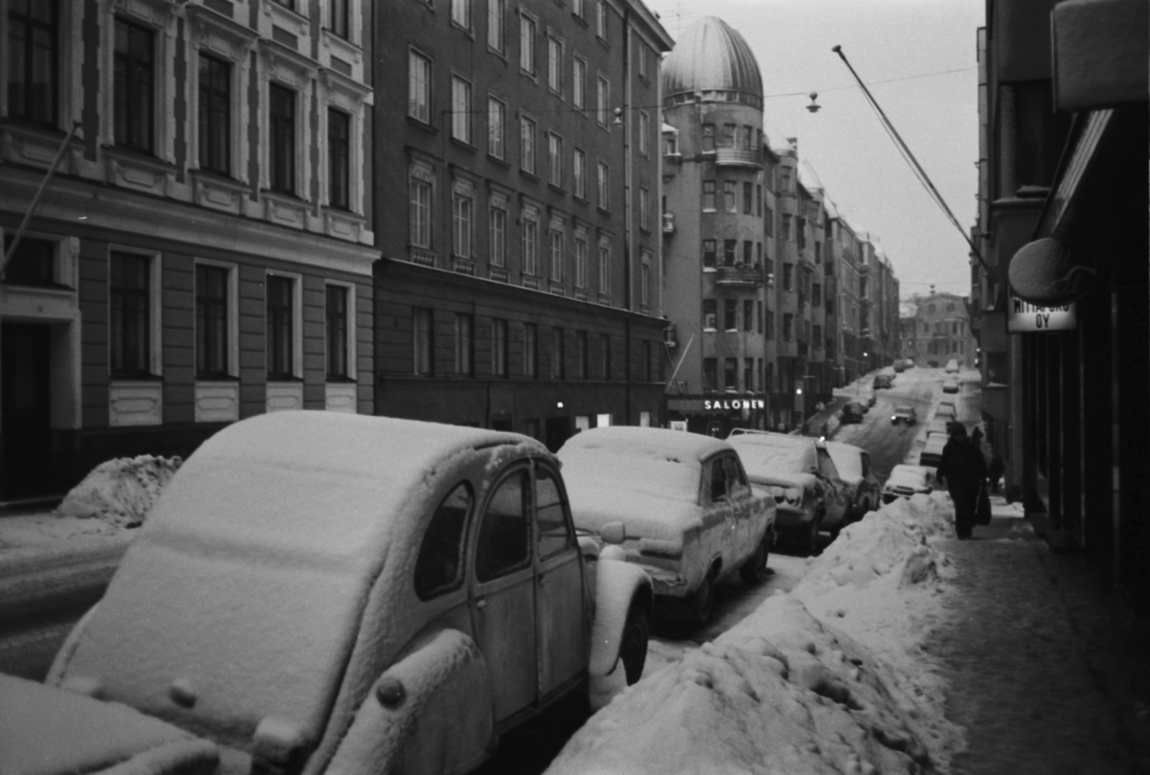 Meritullinkatu. Näkymä lumiselta Meritullinkadulta Maneesikadun kulmasta pohjoiseen. Etualalla kadun laidalla Citroën 2CV, rättisitikka. Taustalla Salosen kaupan valaistu nimi Liisankadun kulmassa.