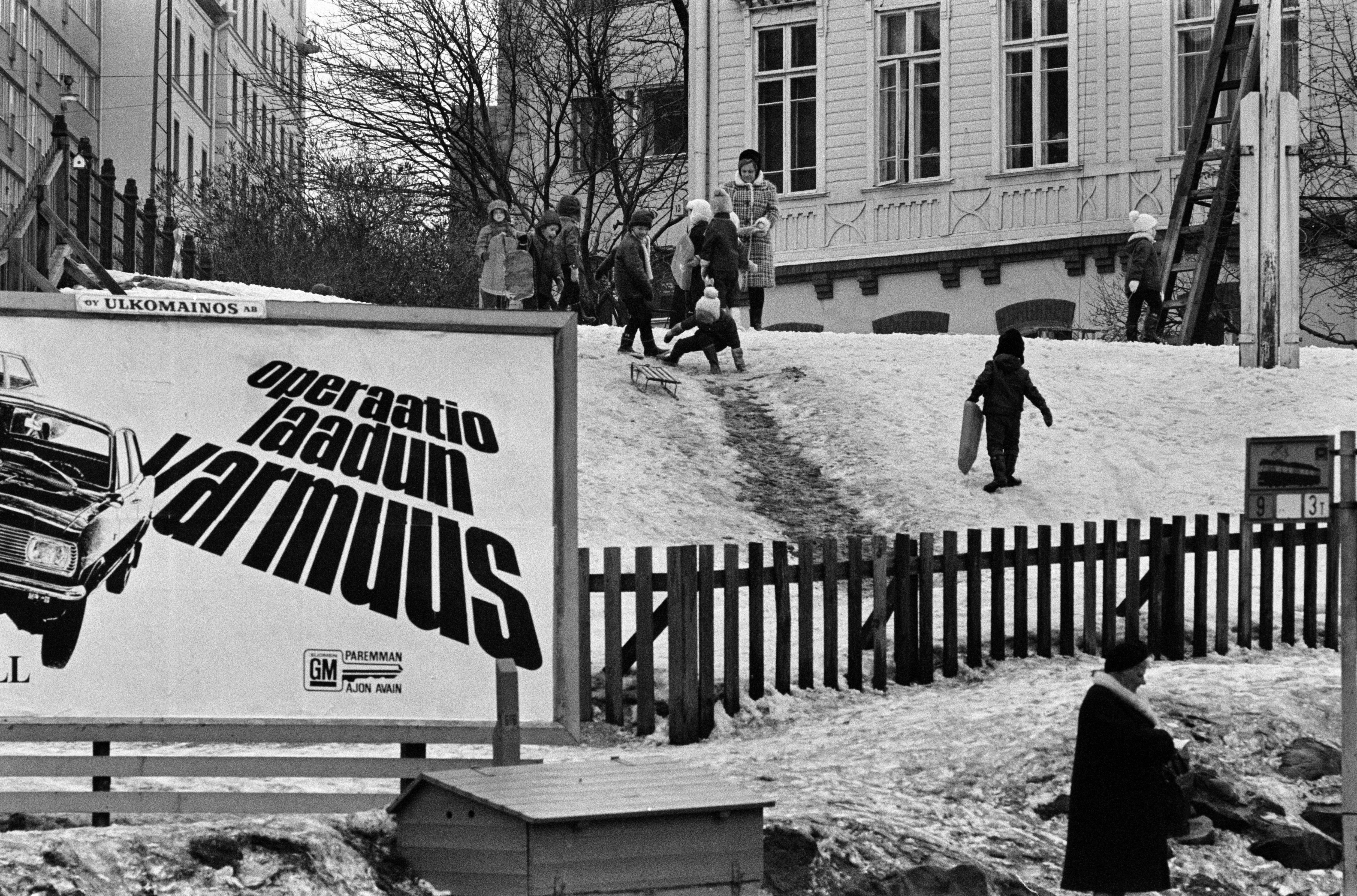 Vauxhall-automainos (Operaatio laadun varmuus) Porthaninkadun ja Siltasaarenkadun kulmassa. Taustalla lapsia laskemassa mäkeä puutalon, Tukholman päiväkodin pihalla. Kevät 1970.