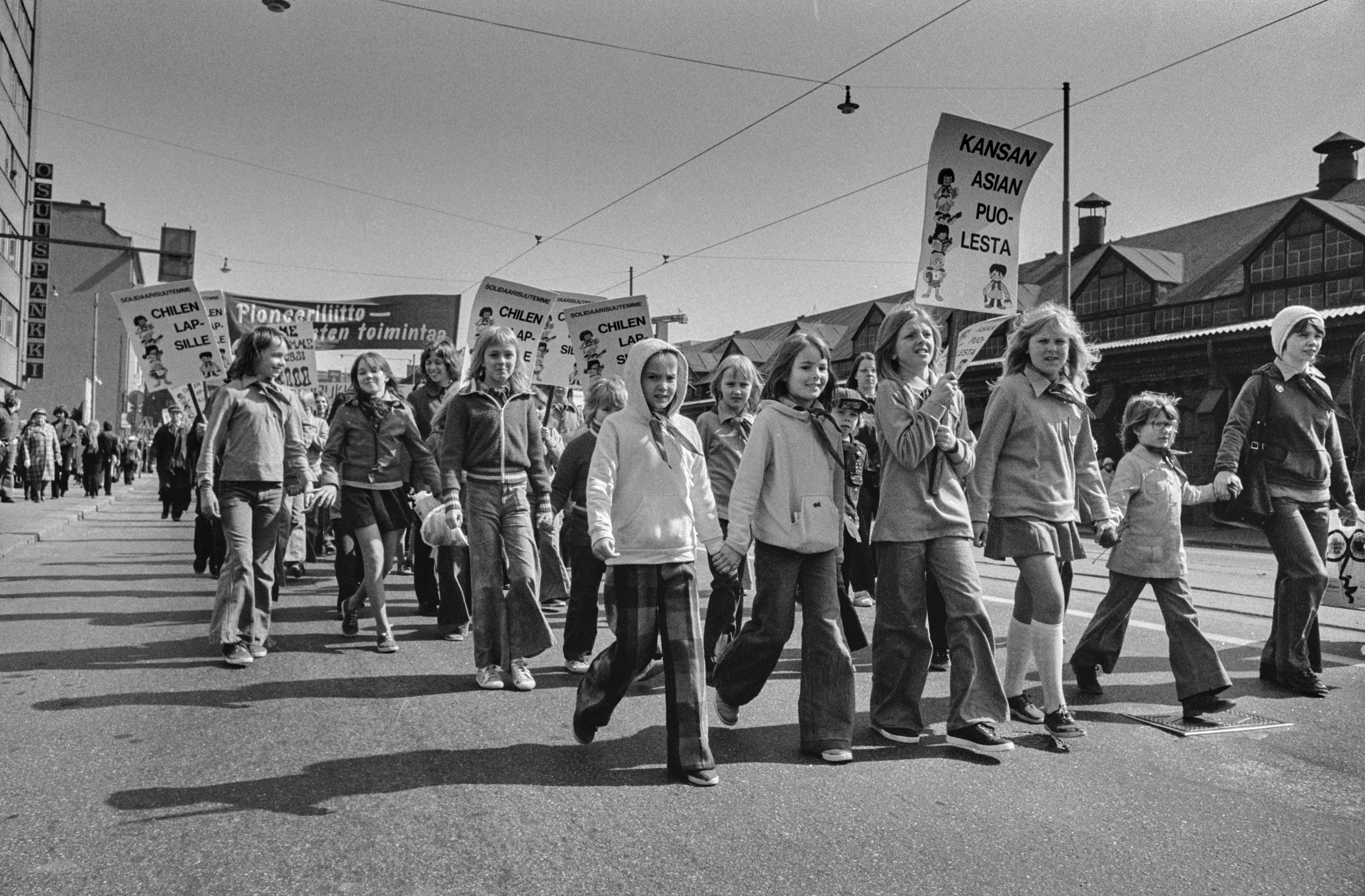 Vappumarssijoita Hämeentiellä. Lapset ja nuoret kantavat kylttejä, joissa tekstit: Solidaarisuutemme Chilen lapsille ja Kansan asian puolesta. Takana Pioneeriliiton banderolli.
