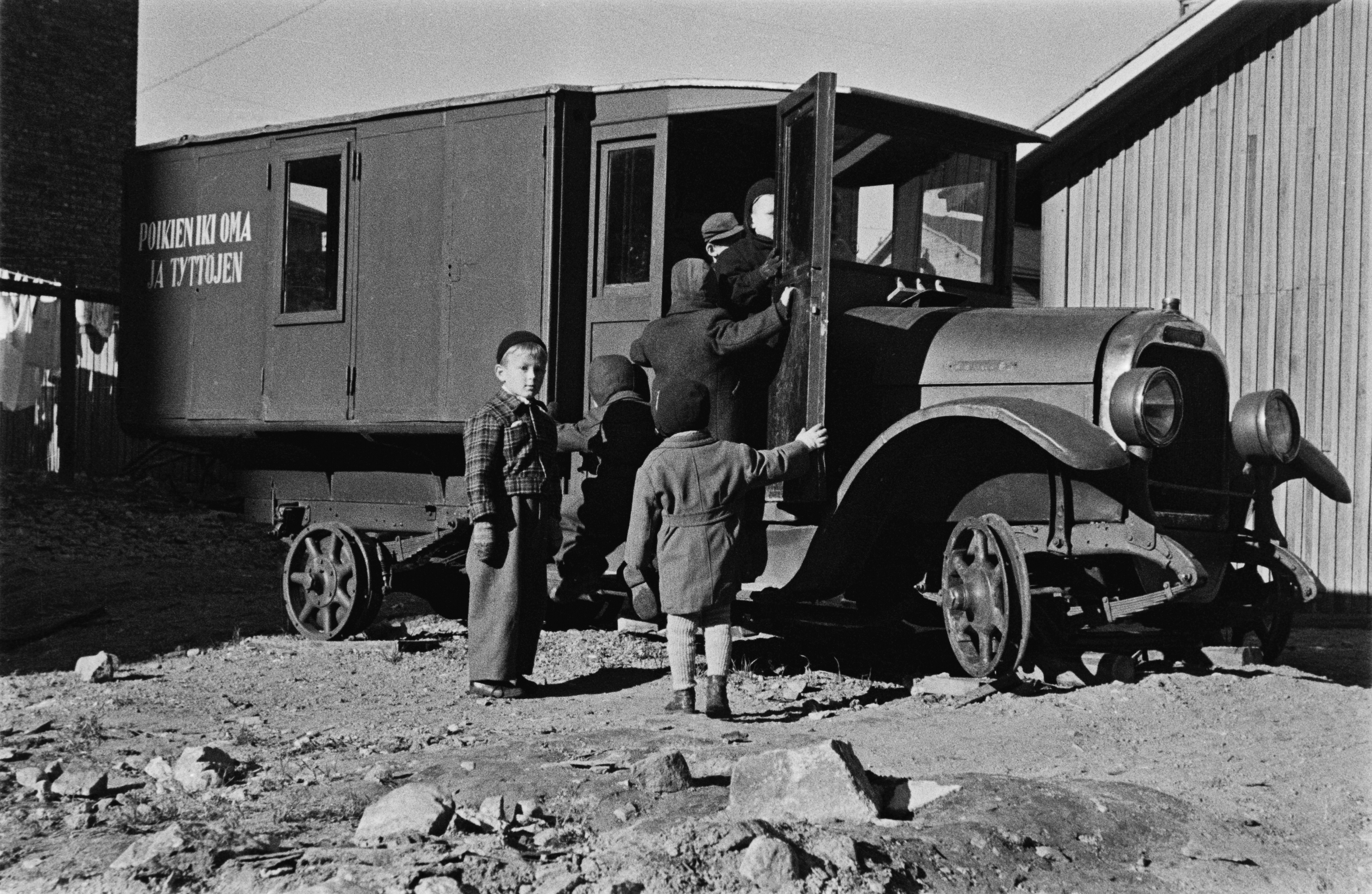 Elannon päiväkodin lapsia leikkimässä Viides linja 4:n pihalla olevan vanhan auton luona. Auto on käytöstä poistettu leipäauto ja siitä on tehty lasten leikkipaikka. Sen kyljessä lukee "poikien ikioma ja tyttöjen".