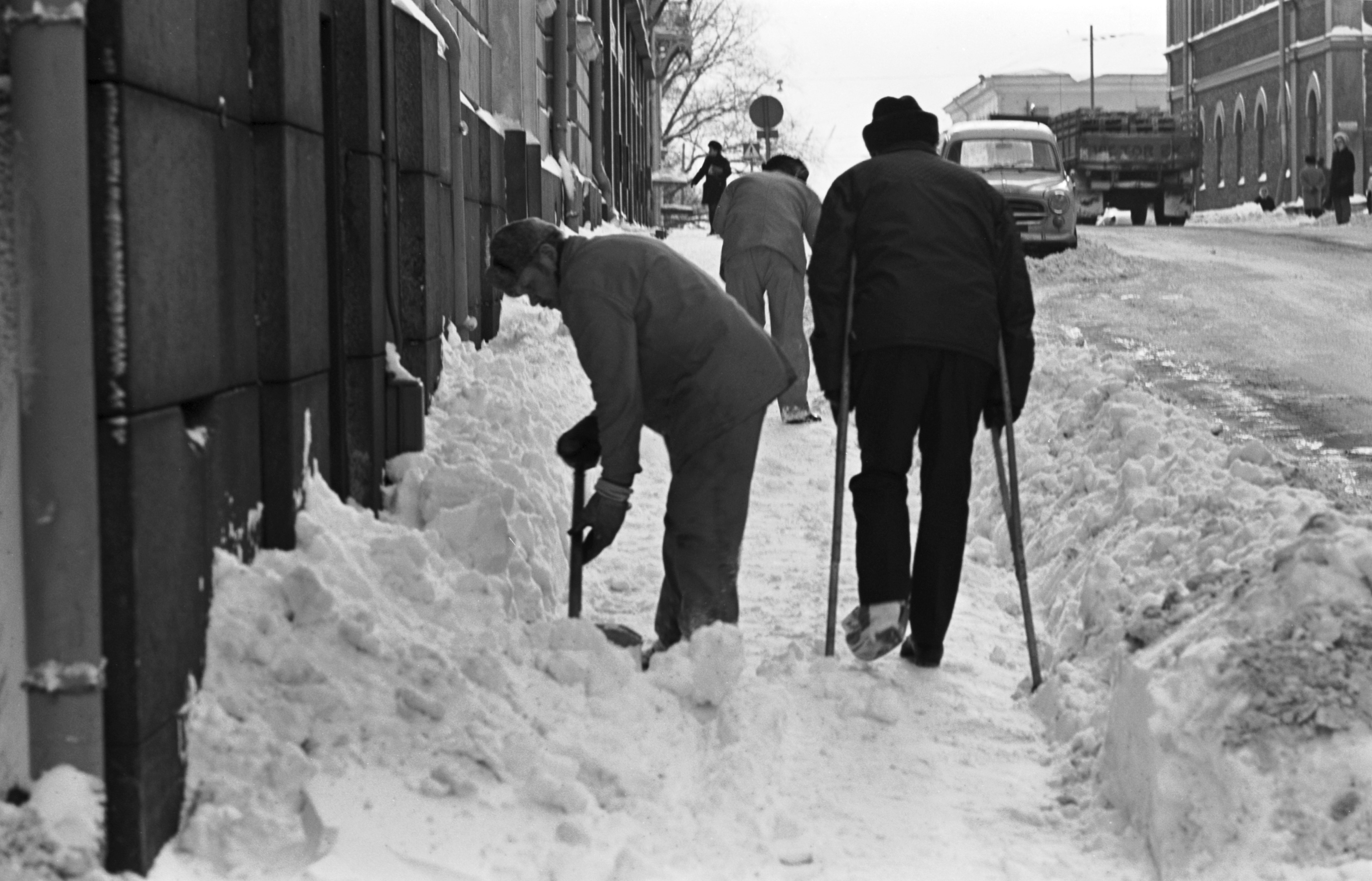 Mariankatu 11. Mies kainalosauvojen kanssa kulkemassa lumisella Mariankadulla, jossa miehiä lapioimassa lunta.