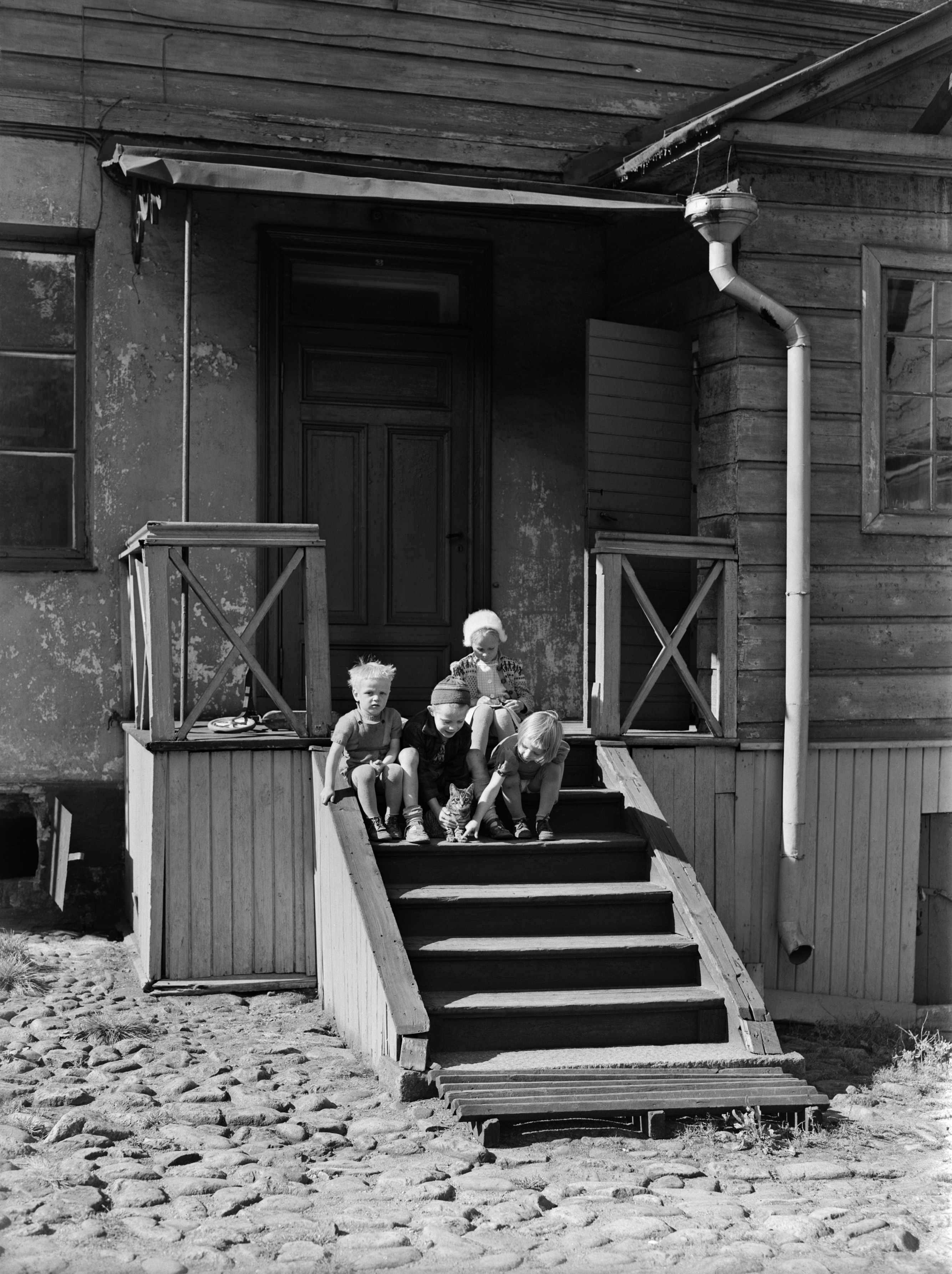 Lapsia ja kissa Snellmaninkatu 21:n päärakennuksen pihanpuolen kuistin portailla. Lasten takana potkulauta.