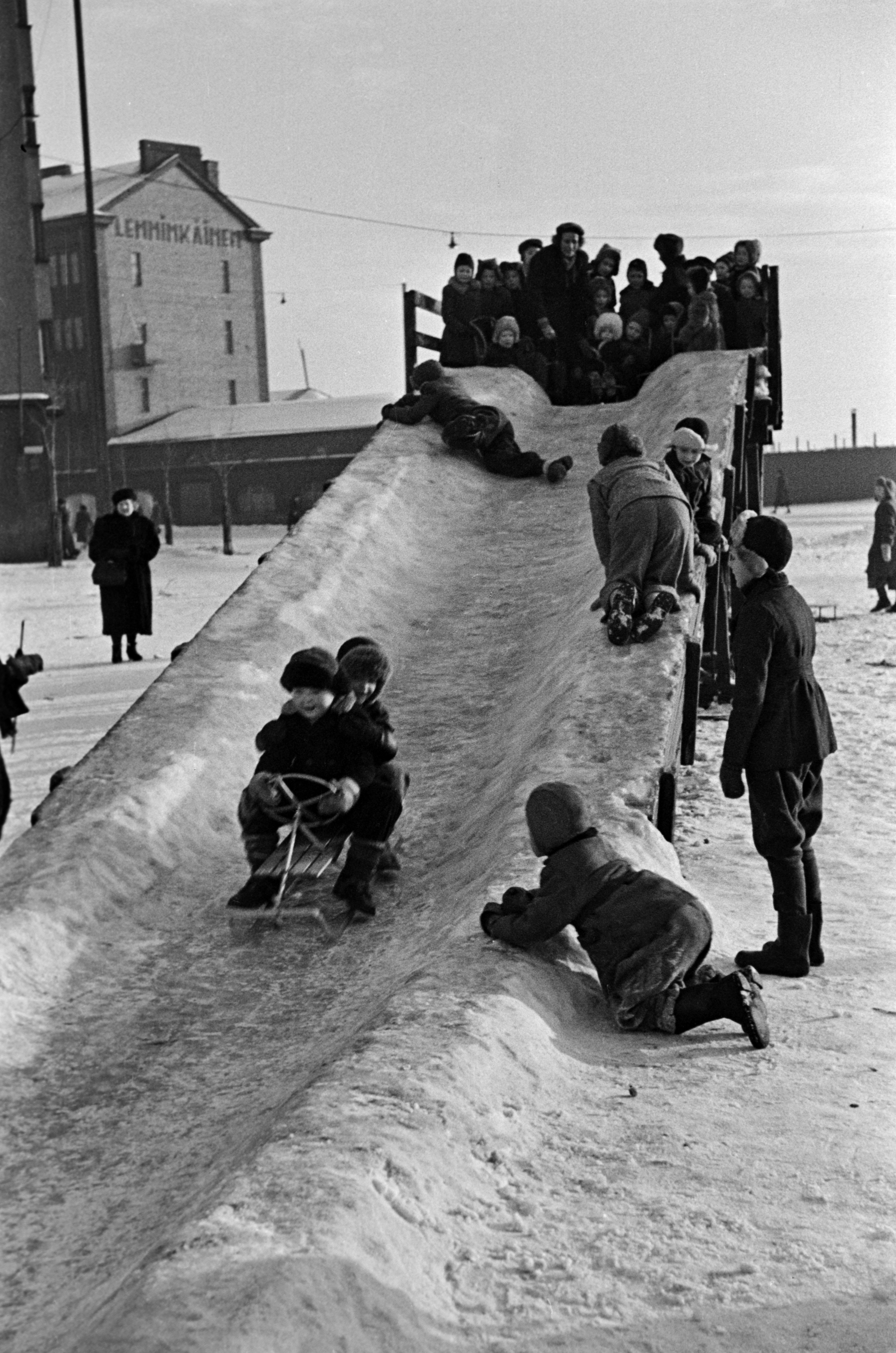Jäämäki ja lapsia laskemassa mäkeä Hakaniementorin kaakkoiskulmassa. Taustalla Hakaniemenkatu 2.