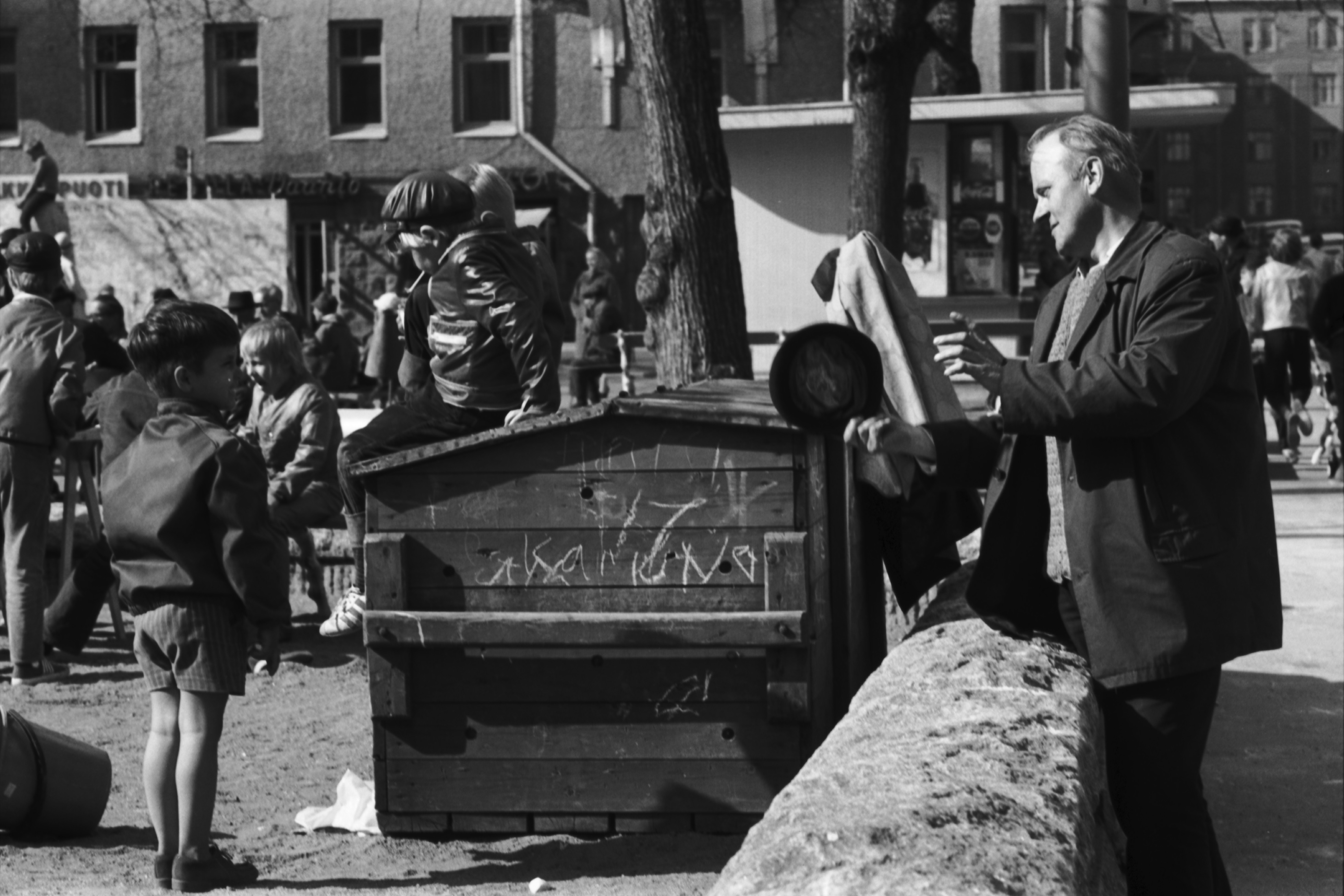 Karhupuisto. Fleminginkatu. Mies lierihattu kädessä Karhupuiston kiviaidan vieressä puistotapahtuman aikaan. Puistossa lapsia.