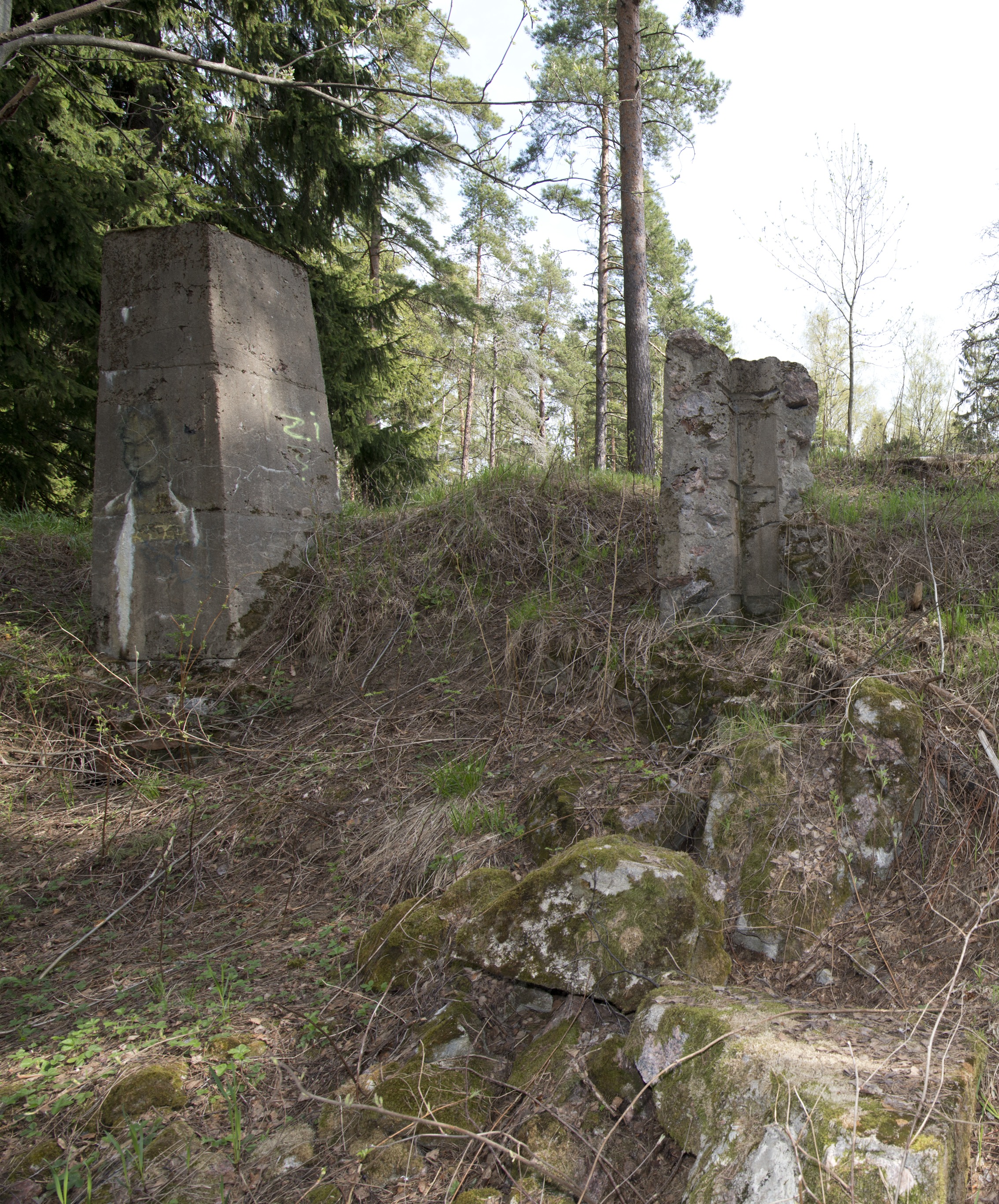 Myllypuro. Ensimmäisen maailmansodan aikainen maalinnoitus, tykkipatteri 65 Myllypurossa. Kuvassa näkyy kaksi pohjoisimman suojahuoneen viidestä betonisesta savupiipusta tai hormista. Vasemmanpuolimmainen hormi on ehyt ja sen kylkeen on maalattu ihmishahmoa esittävä vihertävä graffiti. Oikeanpuolimainen hormi on pahasti vaurioitunut, vain yksi hormin kulmista on enää pystyssä. Kulmissa näkyy painanteina hormia tukeneista vahvikkeista. Kuvaussuuntana pohjoinen.