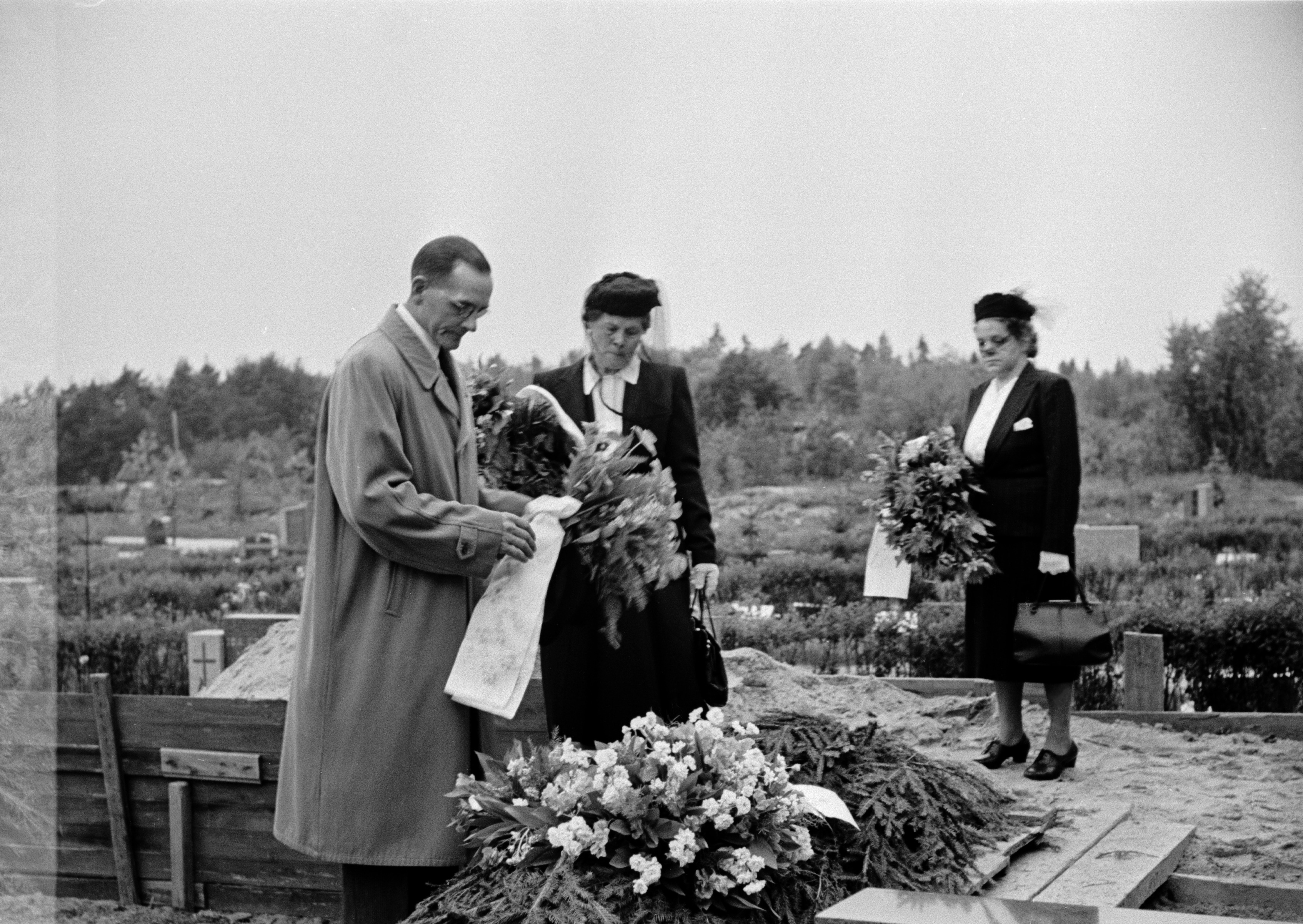 Pohjoinen hautausmaa (=Malmin hautausmaa), hautajaiset. Mies ja kaksi suruhuntuista naista laskevat kukkaseppeleitä haudalle.