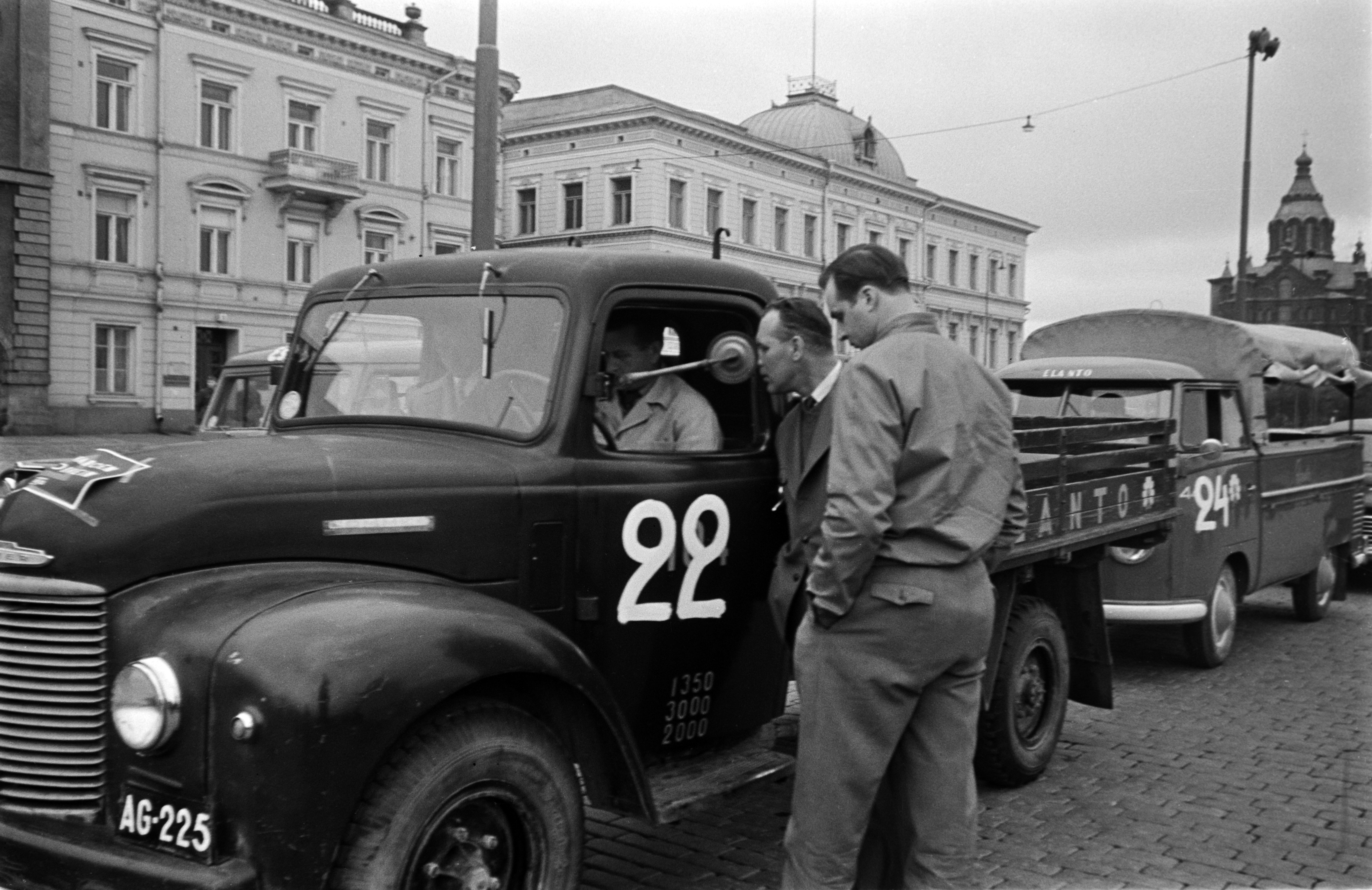 Elannon kuorma-autoja autotapahtumassa. Autojen kylkiin on maalattu numerot ja konepeltiin kiinnitetty teksti "aja varoen, aja oikein". Taustalla Pohjoisesplanadi 5 ja 3 sekä Uspenskin katedraali.