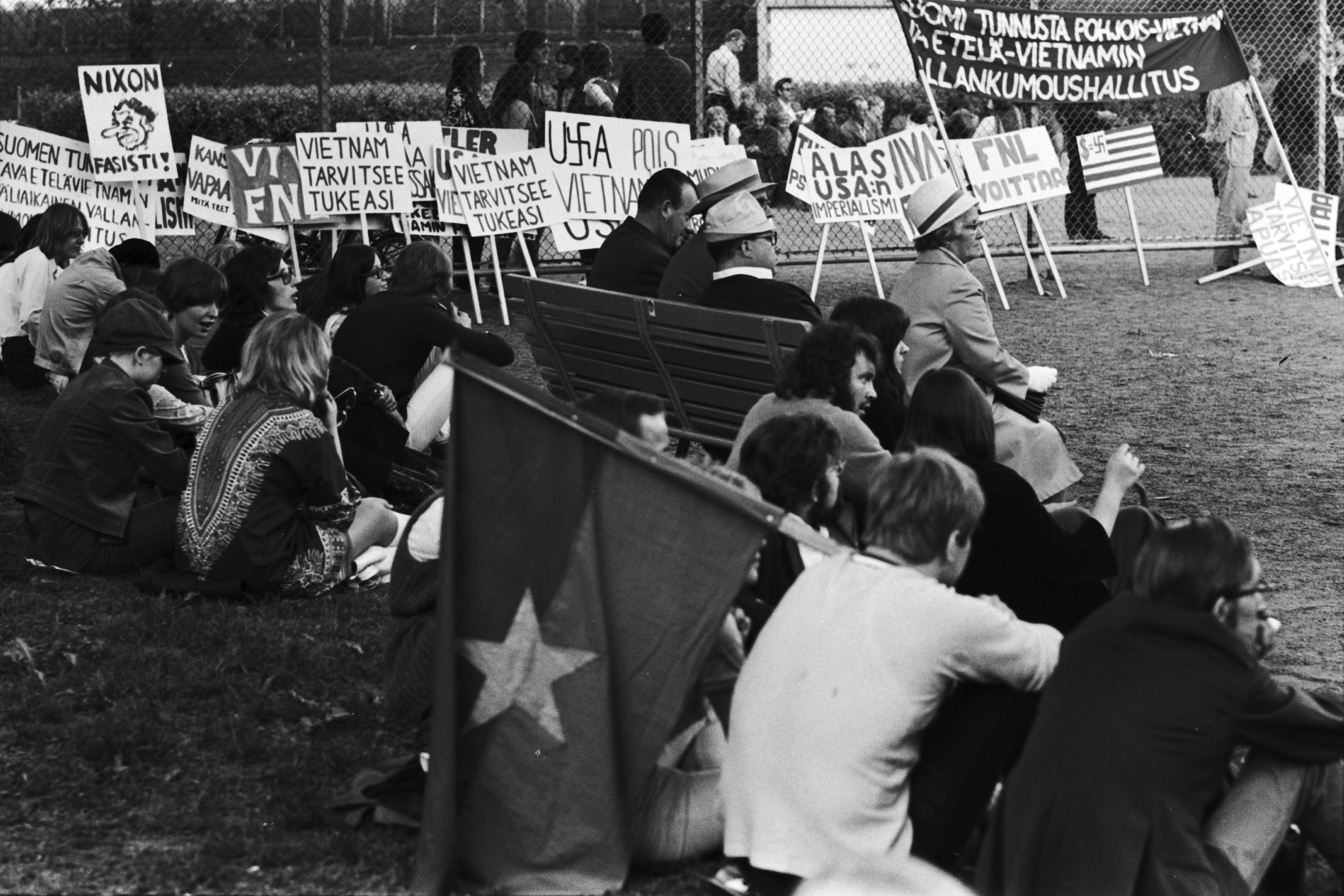 Kaisaniemenpuisto. hmisiä ja aitaa vasten nojallaan olevia kylttejä ja banderolli Vietnamin sodan ja imperialismin vastaisessa mielenosoituksessa Kaisaniemenpuistossa.