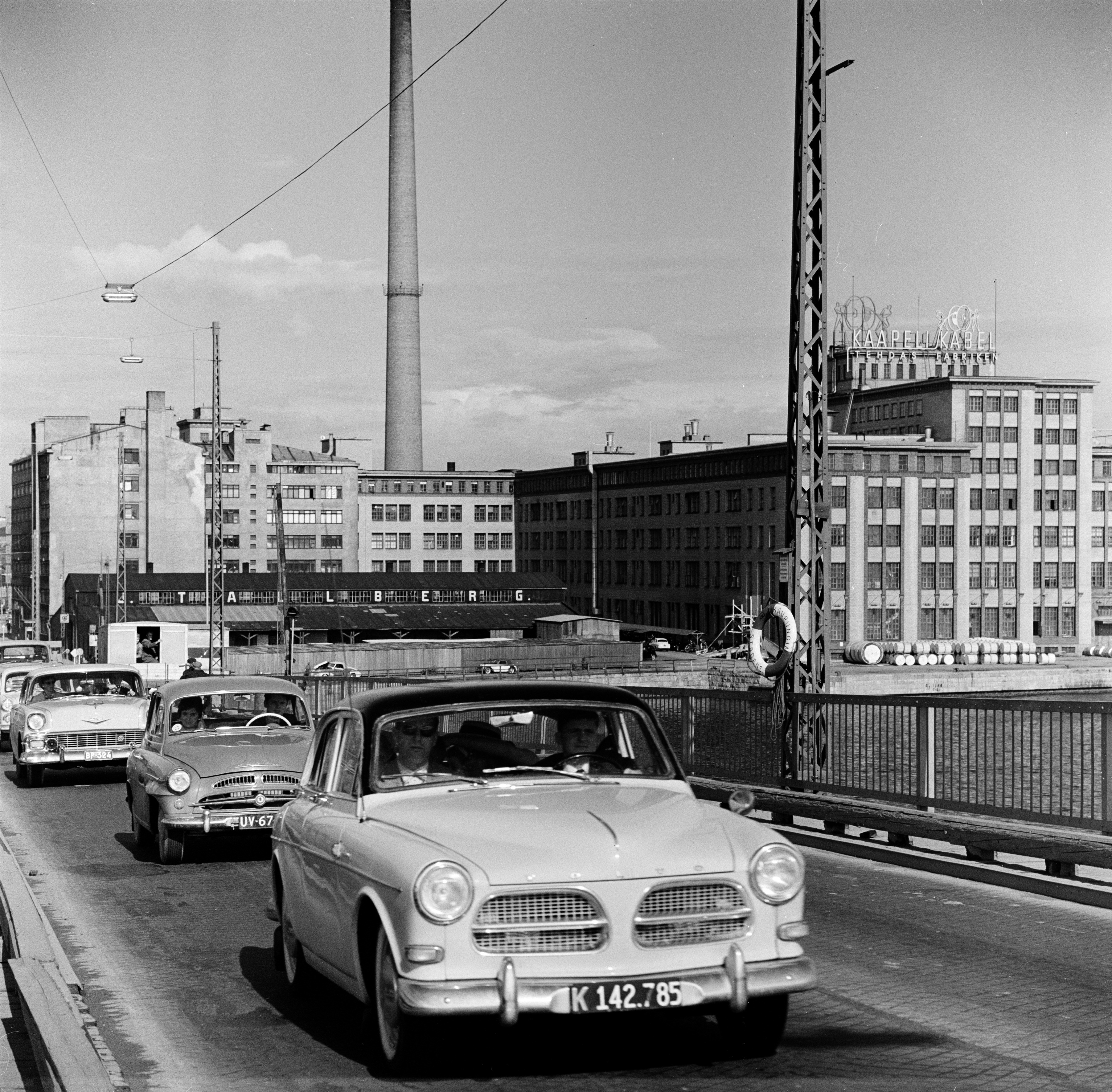 Liikennettä Lauttasaaren sillalla.  Taustalla oikealla Suomen Kaapelitehdas. Myös Julius Tallberg Oy:n varasto näkyy. Ajoneuvot kuvan etualalta taaksepäin lukien: Volvo Amazon, Skoda 440, Chevrolet vm. 1956.