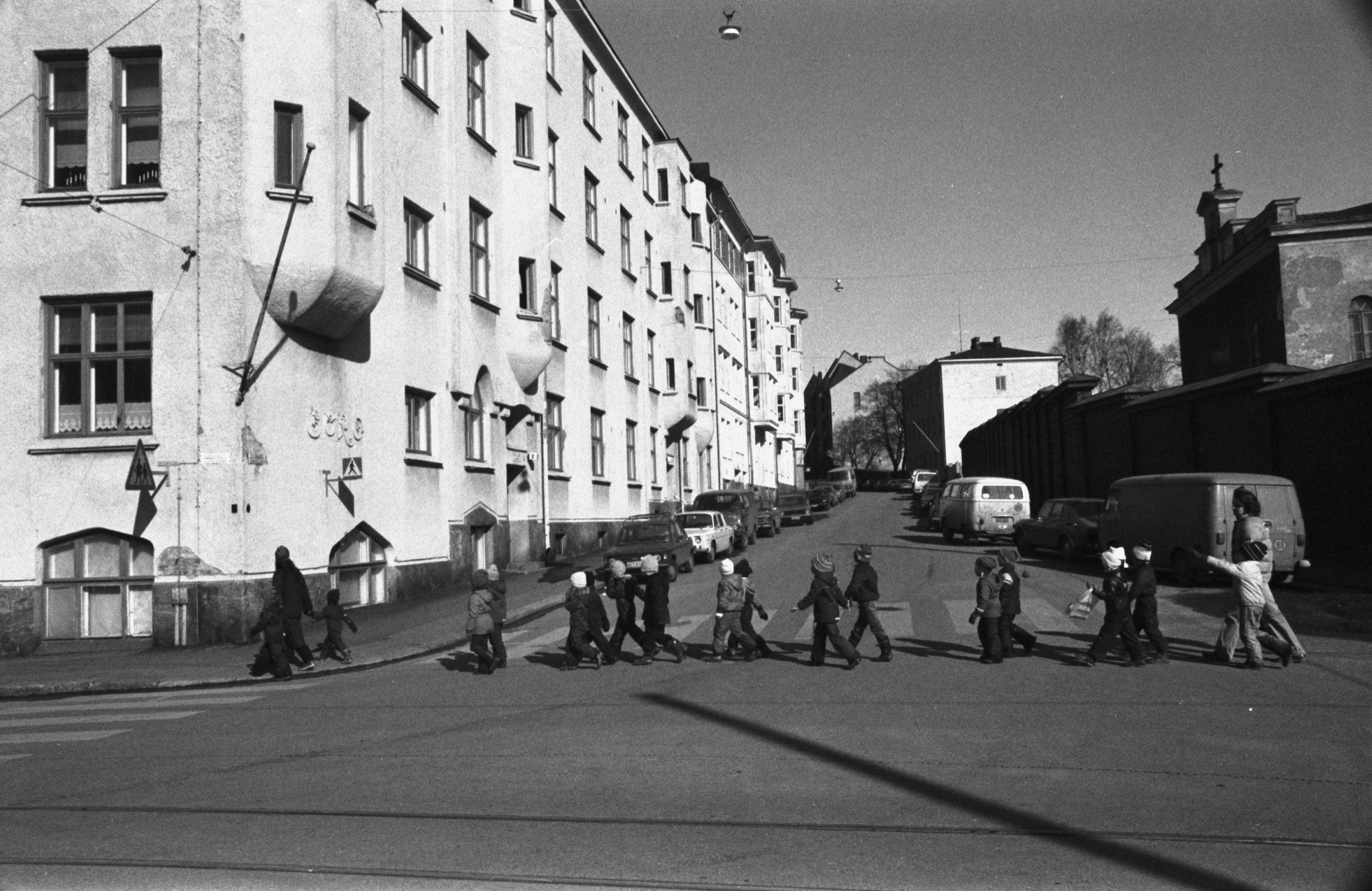 Linnankatu. Vironniemen päiväkodin lapsia ja aikuisia ylittämässä katua Vyökadun kulmassa Katajanokan tutustumiskierroksellaan. Näkymä Linnankadun suunnalta pohjoiseen Vyökadulle. Vasemmalla Linnankatu 7 - Vyökatu 2. Oikealla Katajanokan vankilan muurin takana kappelin pääty.