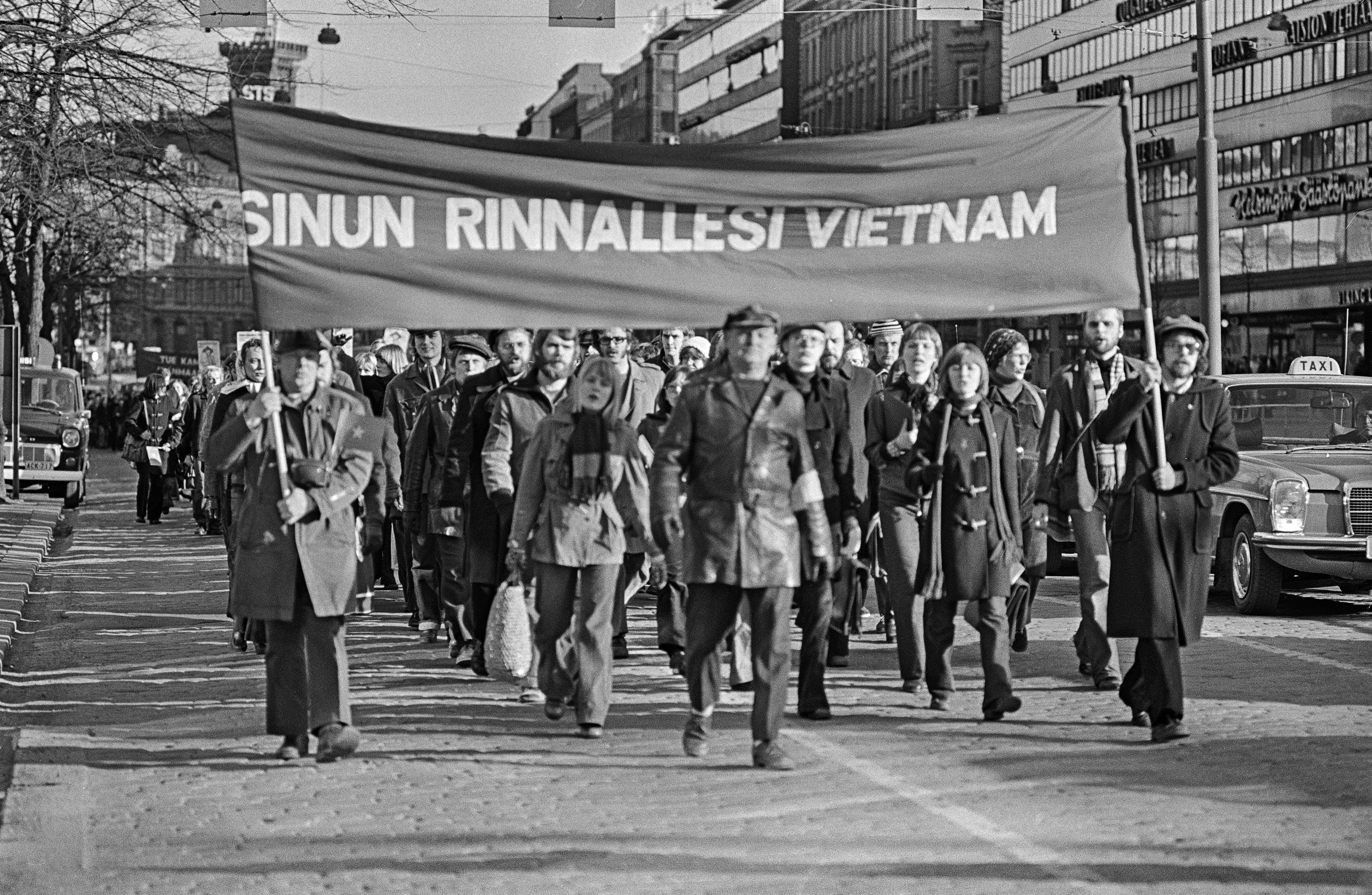 Mielenosoittajia marssimassa ajoradalla Mannerheimintiellä. Mielenosoittajat kantavat banderollia, jossa teksti: Sinun rinnallesi Vietnam. Vasemman puoleisella banderollin kantajalla on mukanaan Vietanamin sosialistisen tasavallan pienoislippu.