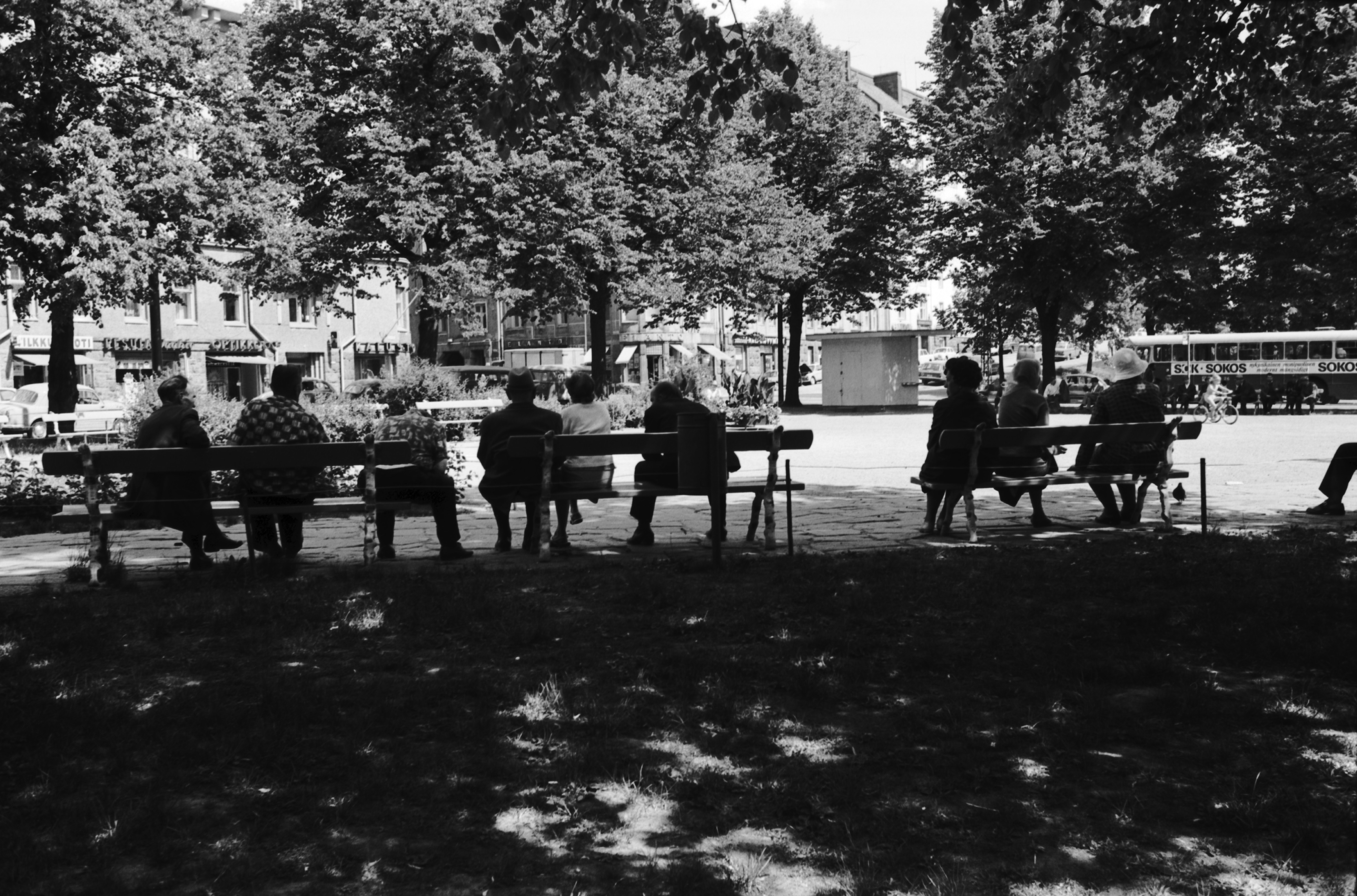 Karhupuisto. Ihmisiä istumassa penkeillä puiden varjossa kesäisen Karhupuiston laidalla. Näkymä Viidenneltä linjalta Agricolankadun ja Fleminginkadun risteyksen suuntaan. Taustalla lippakioski.