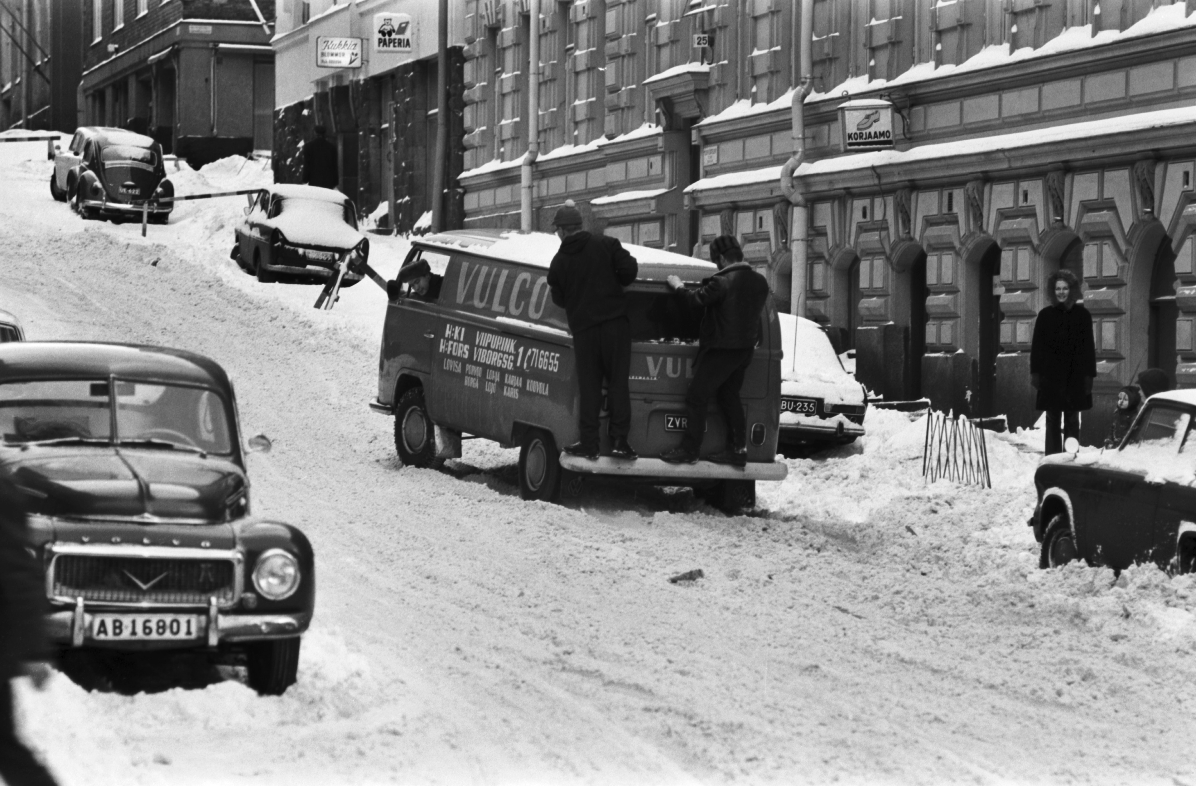 Meritullinkatu 25 - 27. Kaksi miestä seisomassa painona Volkswagen -pakettiauton peräpuskurilla lumisella Meritullinkadulla. Näkymä Liisankadun kulmasta Kruununhaankadun suuntaan.
