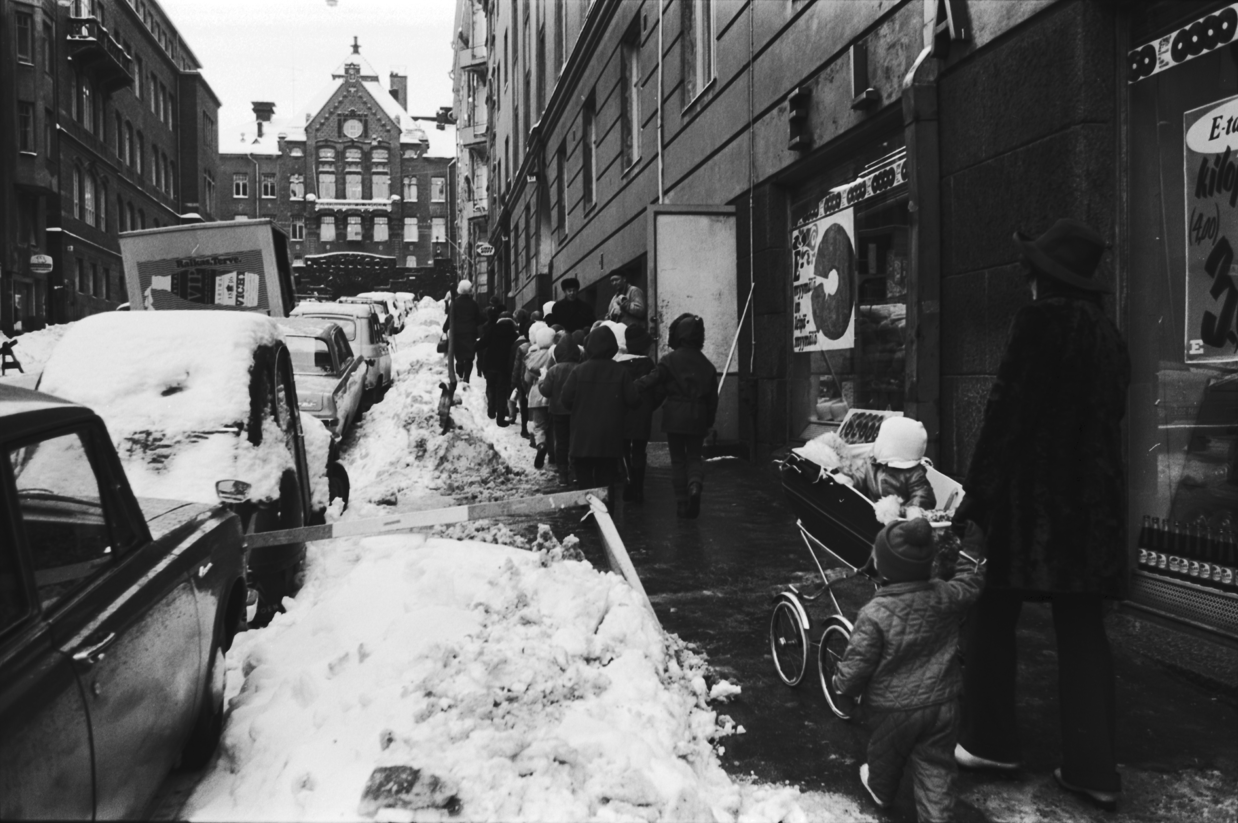 Kruununhaka, Snellmaninkatu. Jonossa kulkevia lapsia sekä lastenvaunuja työntävä nainen kävelemässä ylös lumikinoksen kaventamaa Snellmaninkatua Liisankadun kulmasta Oikokadulle päin. Oikealla Elannon myymälä ja taaempana lihakaupan kyltti.