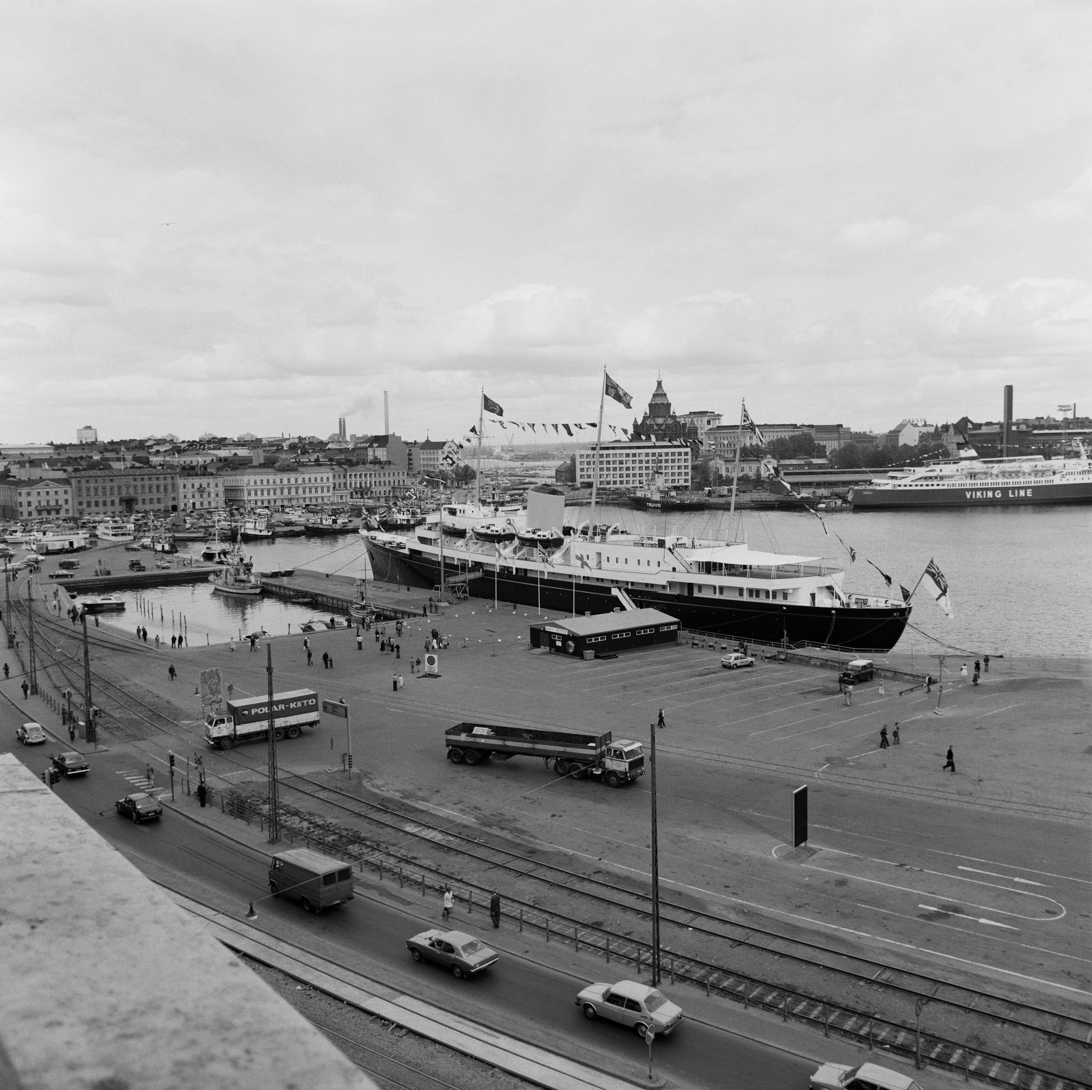 HMY Britannia Pakkahuoneenlaiturissa, Englannin kuningatar Elisabeth II:n valtiovierailu. Aluksen vasemmalle puolelle jää Vironallas, taustalla on Kauppatori takanaan Pohjois-Esplanadin taloja. Niistä oikealla on Enso Gutzeitin pääkonttori Katajanokalla takanaan Uspenskin katedraali. Kuvattu mahdollisesti Palace-hotellista.