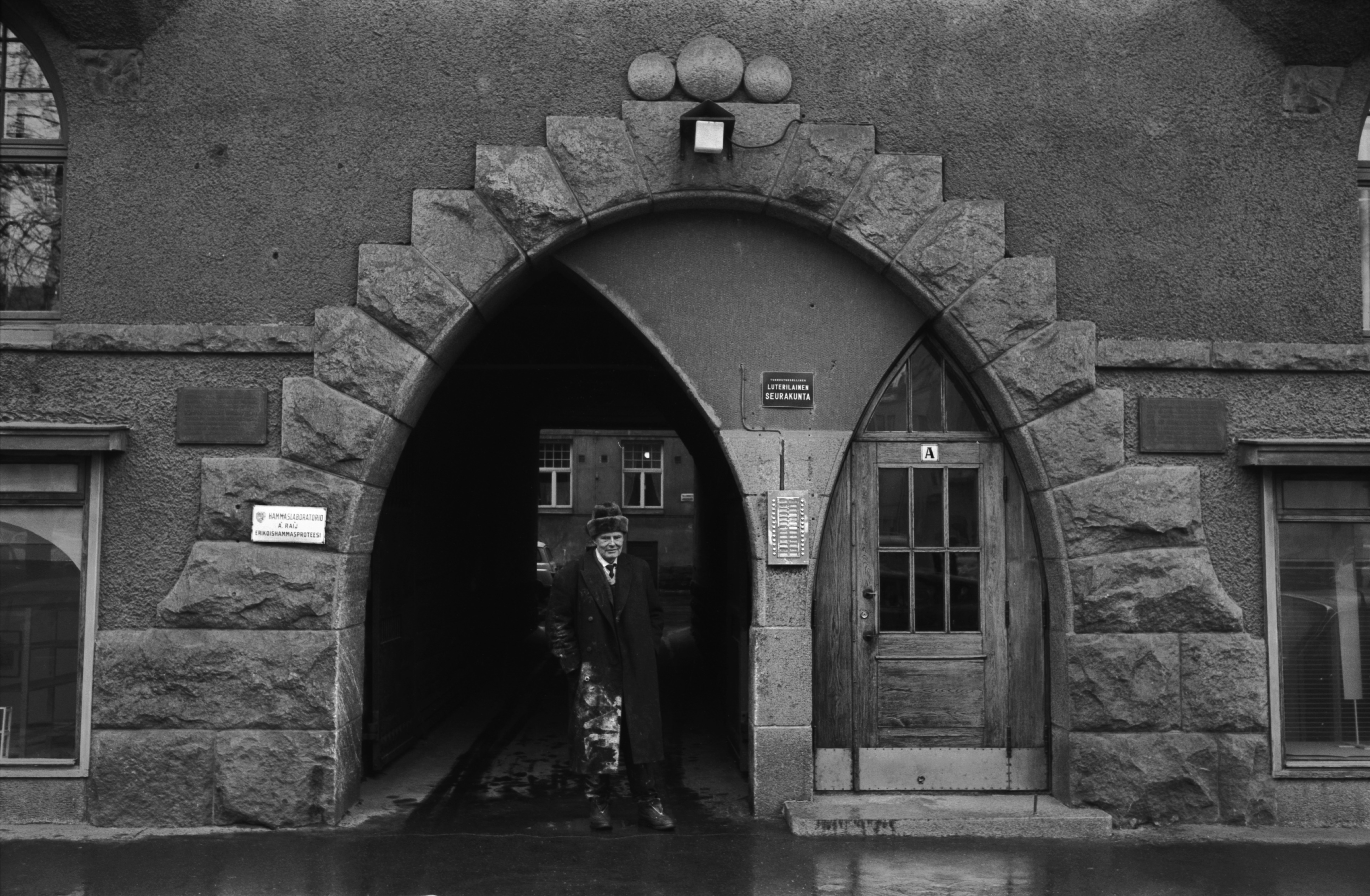 Fabianinkatu 13. Mies, jonka ulsterissa valkoista maalia(?) seisomassa sateisella Fabianinkadulla jugendtalo Torilinnan porttikäytävän edustalla Kasarmitorin laidalla.
