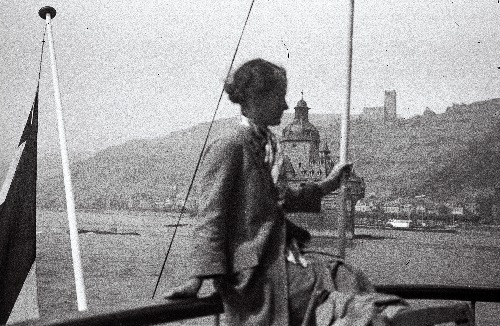 Lore von Krusenstiern nõjatumas laeva käsipuule. Taustal jõeäärne linnake ning loss-kindlus.