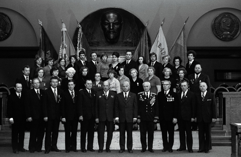 Eesti NSV Kaubandusettevõtete ja organisatsioonide töötajate, sotsialistliku võistluse võitjate pildistamine revolutsiooni-, lahingu - ja töökuulsuse lippude juures Eesti NSV Ministrite Nõukogu Valges saalis seoses Suure Sotsialistliku Oktoobrirevolutsiooni 60. aastapäeva tähistamisega. Tallinna Kaubamaja direktor Toivo Rüekenberg, Tallinna 1. Toitlustustrusti kohviku Tallinn direktor, Eesti NSV teeneline kaubandustöötaja Arnold Rünkaru, Tallinna 2. Toitlustustrusti restorani Tallinn-Balti tootmisalajuhataja Emmeline-Hildegard Vint, Tallinna Kaubamaja sektsioonijuhataja Helju Plaan,  jätkub: Tallinna Tööstuskaubastu kaupluse nr. 78 filiaali müüja Marika Prits, Tallinna 1. Toitlustustrusti restorani Kännu-Kukk kokk Tatjana Ratnikova, Viljandi Kaubastu kaupluse Galanterii sektsioonijuhataja Sirje Reier, Viljandi Kaubastu restorani Viljandi kaubatundja Maire Sarri, Tartu Toitlustustrusti kohviku nr. 20 filiaali juhataja Uudo Serg, Tallinna 2. Toitlustustrusti söökla