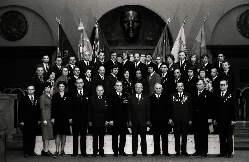 Masina- ja aparaadiehitustööstuse töötajate, sotsialistliku võistluse võitjate pildistamine revolutsiooni-, lahingu- ja töökuulsuse lippude juures Eesti NSV Ministrite Nõukogu valges saalis seoses Suure Sotsialistliku Oktoobrirevolutsiooni 60. aastapäeva tähistamisega, Lauristini nimelise Tallinna Masinatehase lukksepp, sotsialistliku töö kangelane Hans Liblik, personaalpensionär, sotsialistliku töö kangelane Alfred Valdov, puurijad Hilja Päer ja Valeri Fomenkov, lukksepad Nikolai Karpin ja Mihhail Golubkov, katelsepp Viktor Rudakov, direktor Viljar Veskiväli; tootmiskoondise Talleks treial Valeri Liiv, Paide filiaali ülem Vil Tago, vormija Viktor Agafonov, treial Aulis Ventsel, lukksepp Heino Piir, peadirektor Endel Innos;   jätkub: Tartu Aparaadiehituse Tehase treialid Pavel Gorjunov ja Tiit Silm, elektrikeevitaja Arnold Laur, lukksepp-monteerijad Maria Tiideman ja Ljudmila Fenina, trükiskeemide valmistaja Helju-Mare Terentjeva, lukksepp Harri Maasing; tehase Ilm